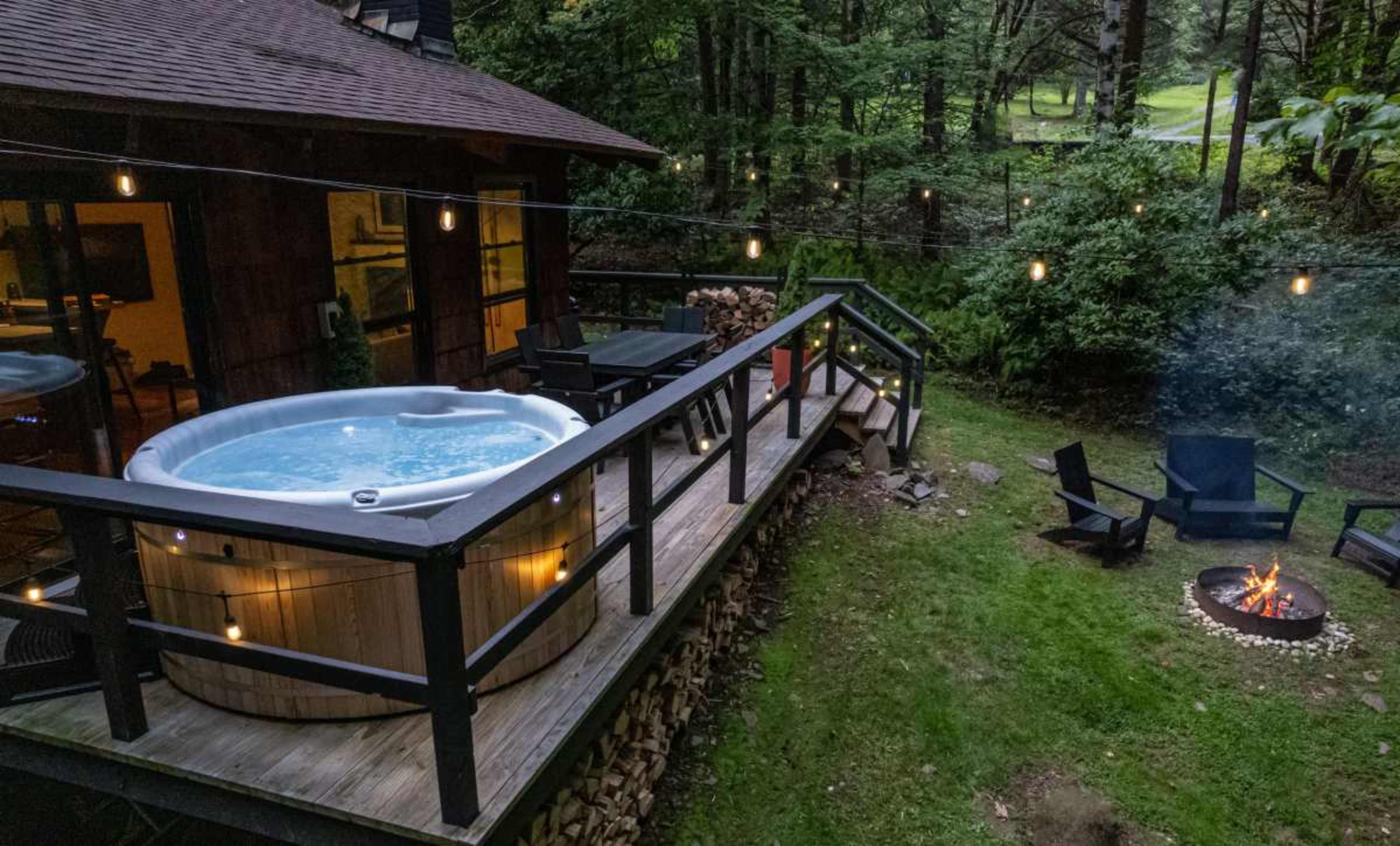 A wooden deck features a hot tub and a seating area with chairs around a fire pit, surrounded by trees.