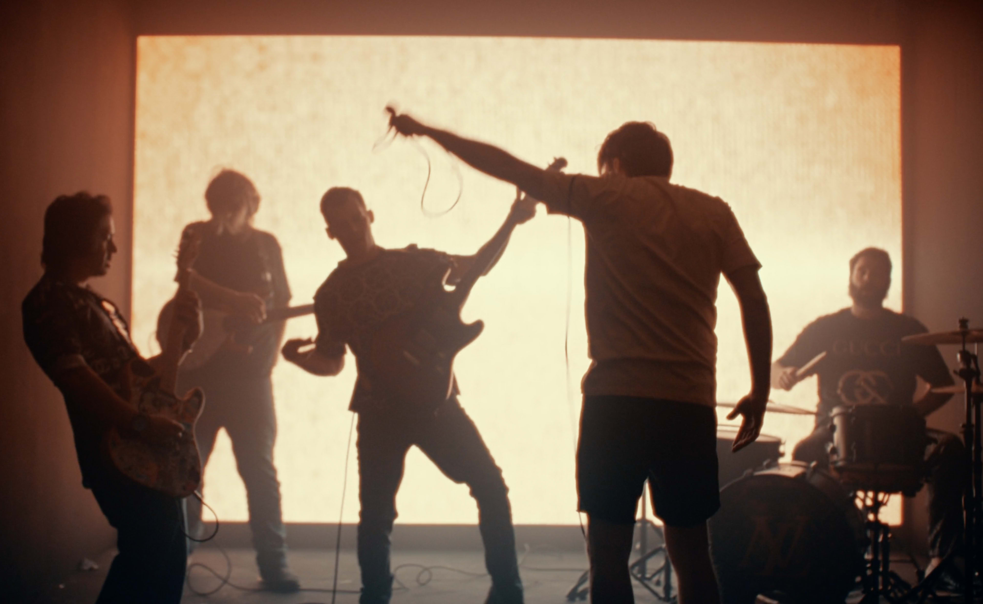 A band performs on stage in front of a bright backdrop, with members playing instruments and singing.