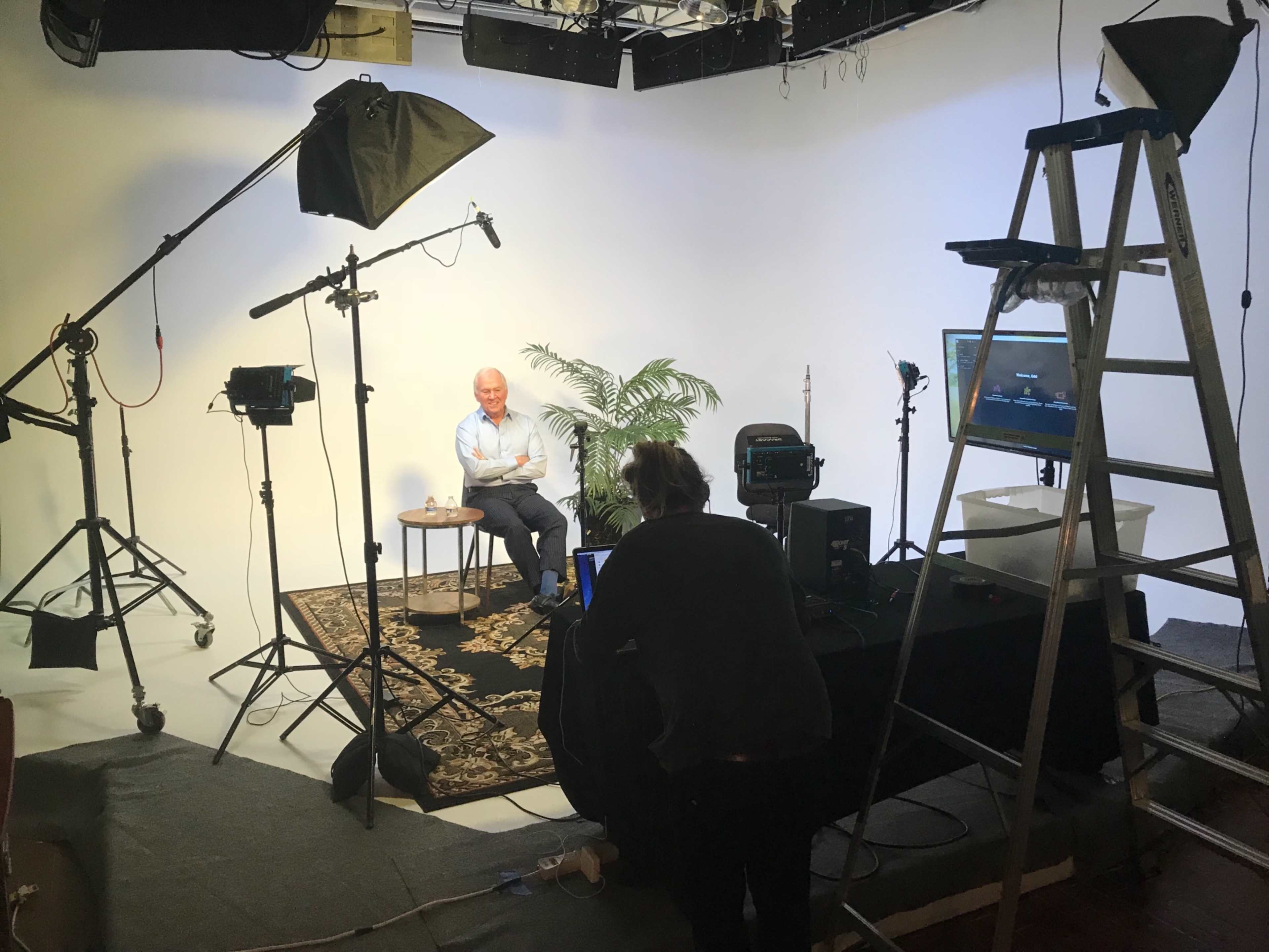 A person sits on a chair in a studio setup with lighting equipment, cameras, and a ladder positioned nearby.