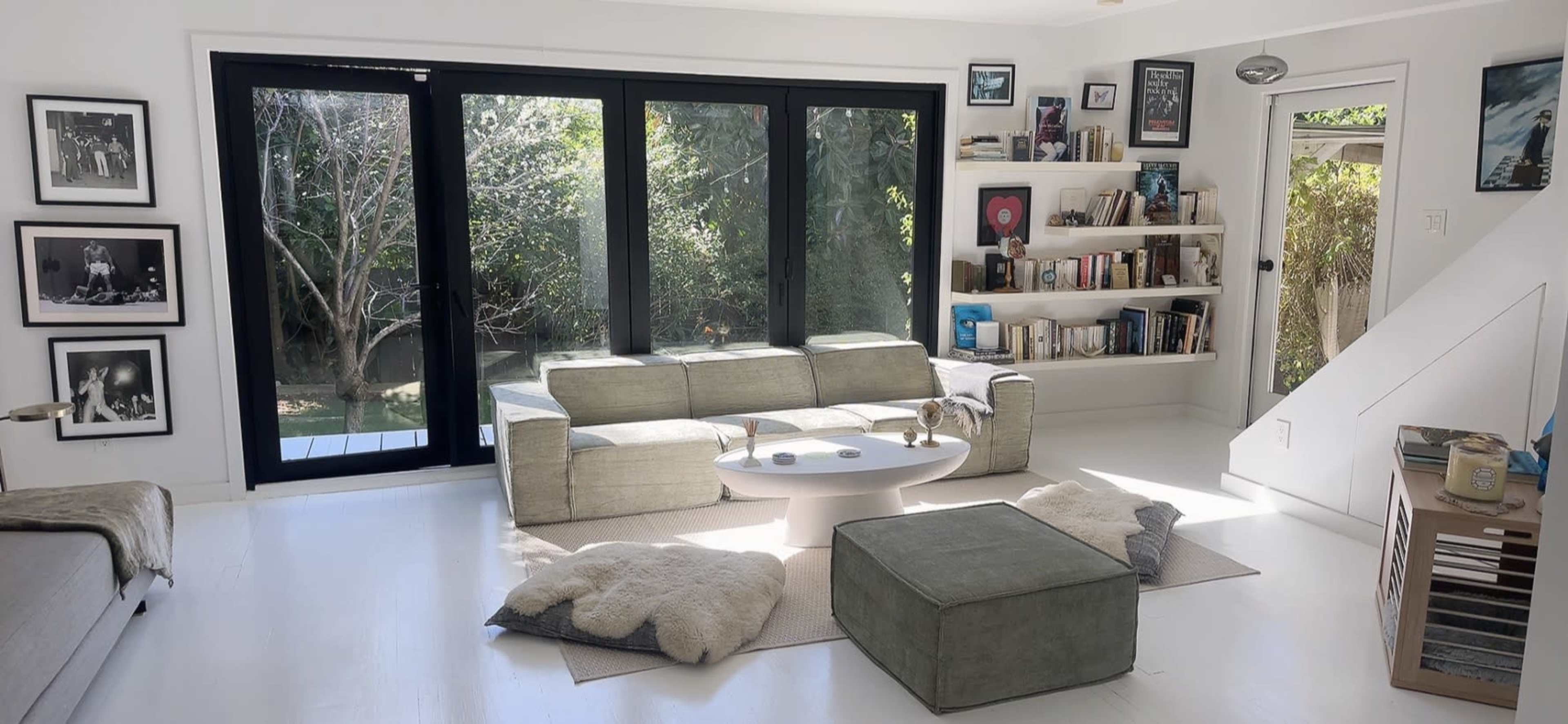 A modern living room features a large window with views of greenery, a grey sofa, a round coffee table, and a bookshelf filled with books and decor.