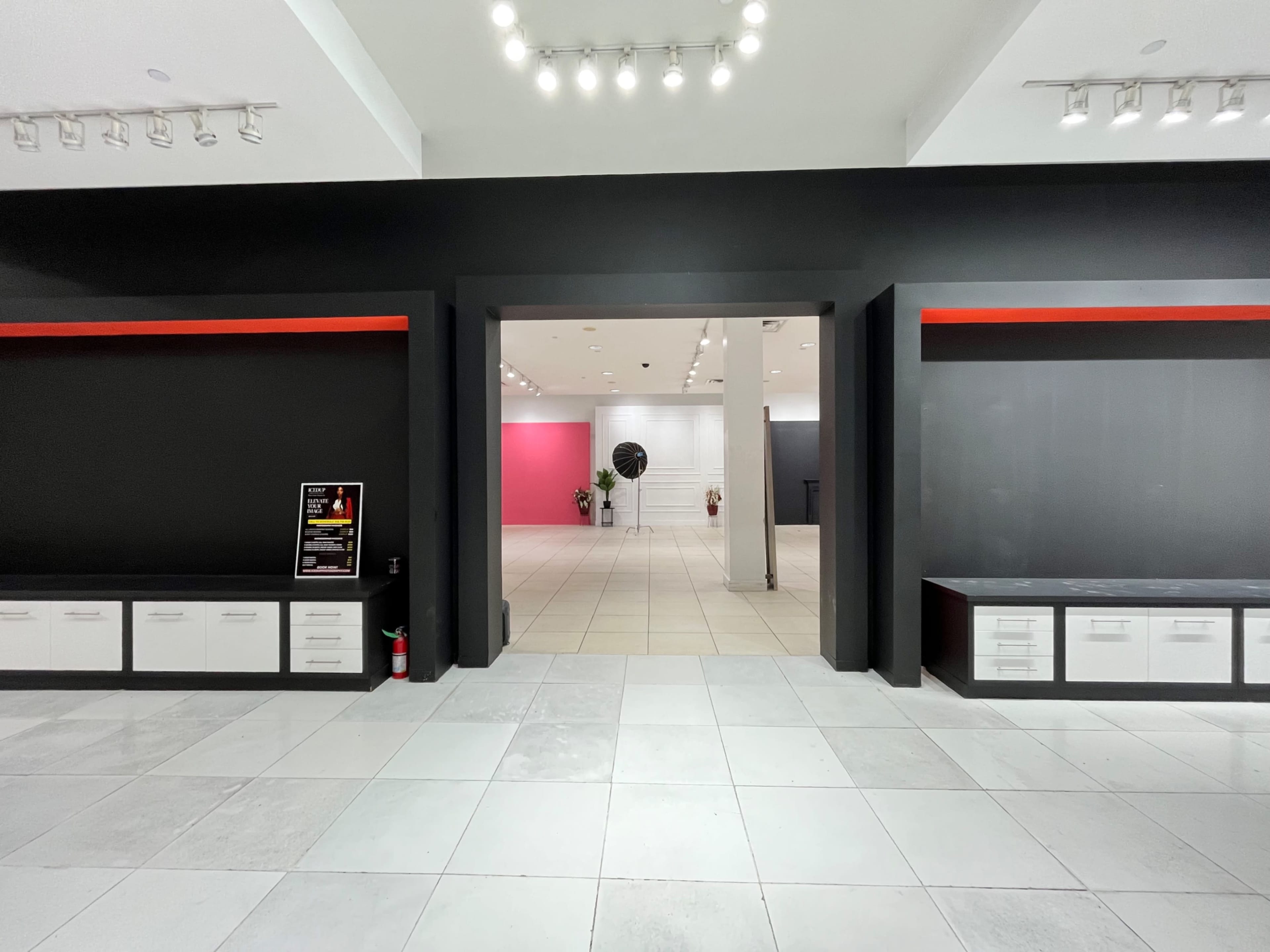 The image shows a spacious, modern interior with black walls, white tile flooring, and decorative red accents, leading to a bright area with pink and gray walls.
