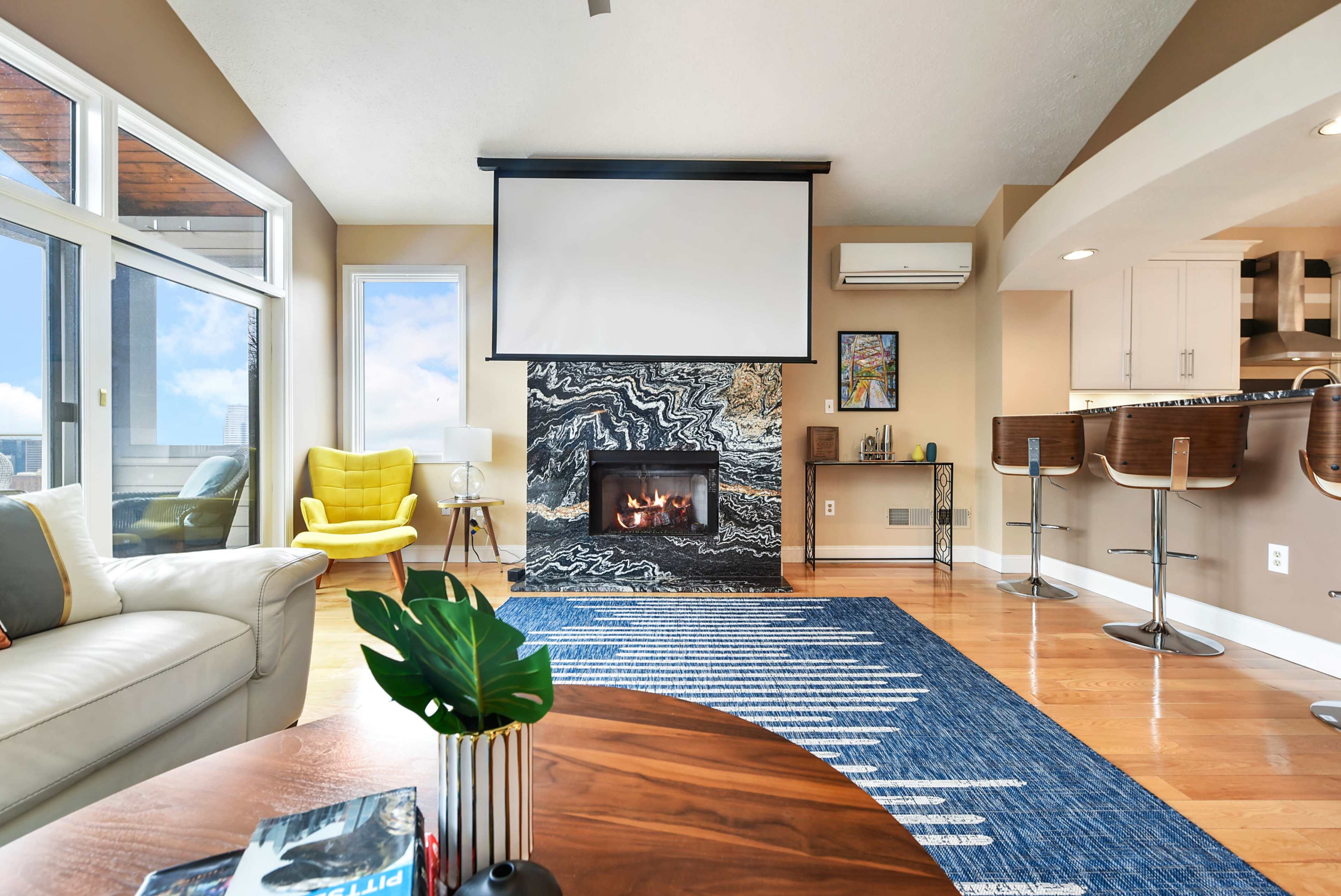 A modern living room features a marbled fireplace, a large screen mounted above it, and seating areas with vibrant yellow and white accents.