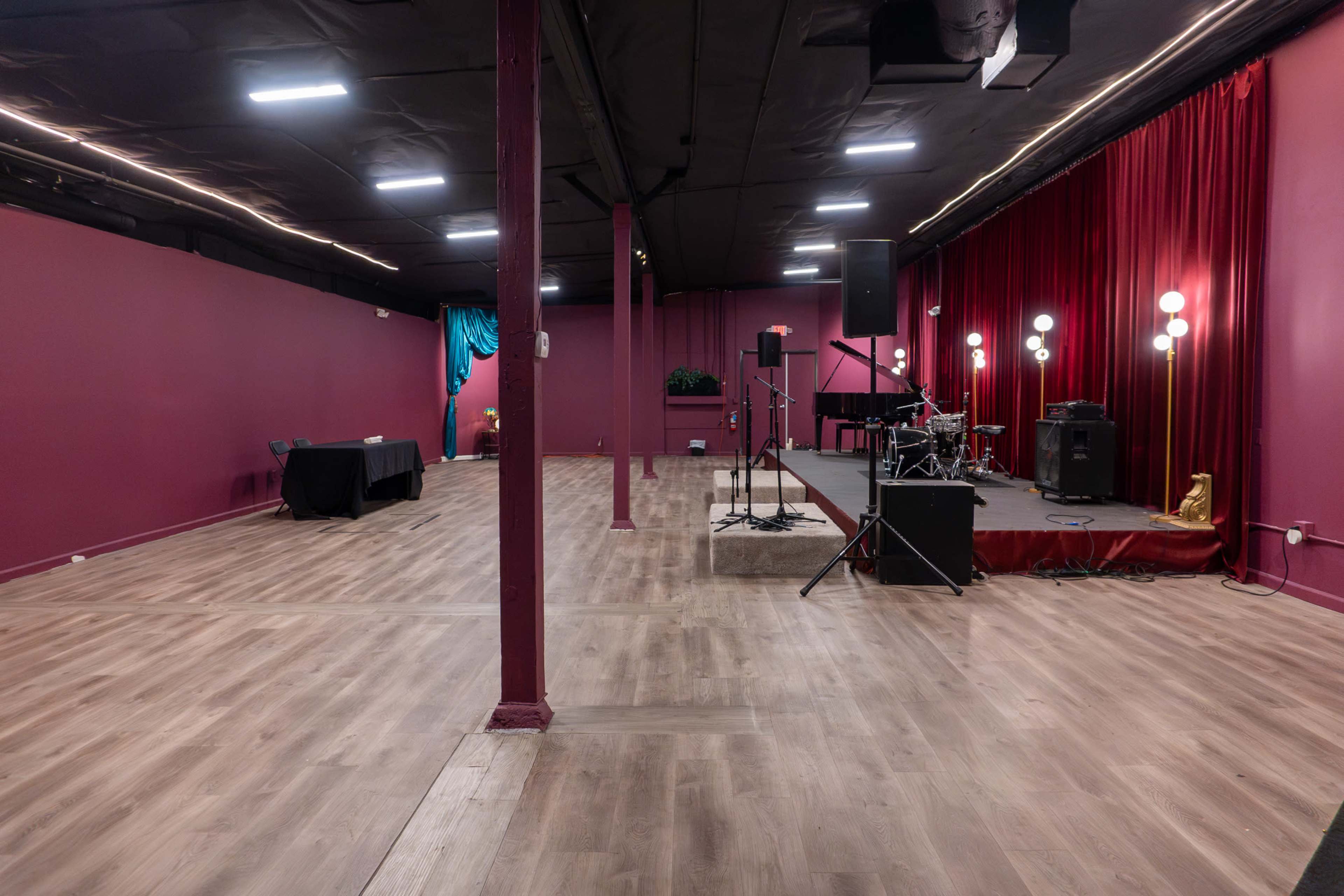 The image shows an empty performance space with wooden flooring, a stage set with musical equipment, and a backdrop of red curtains.