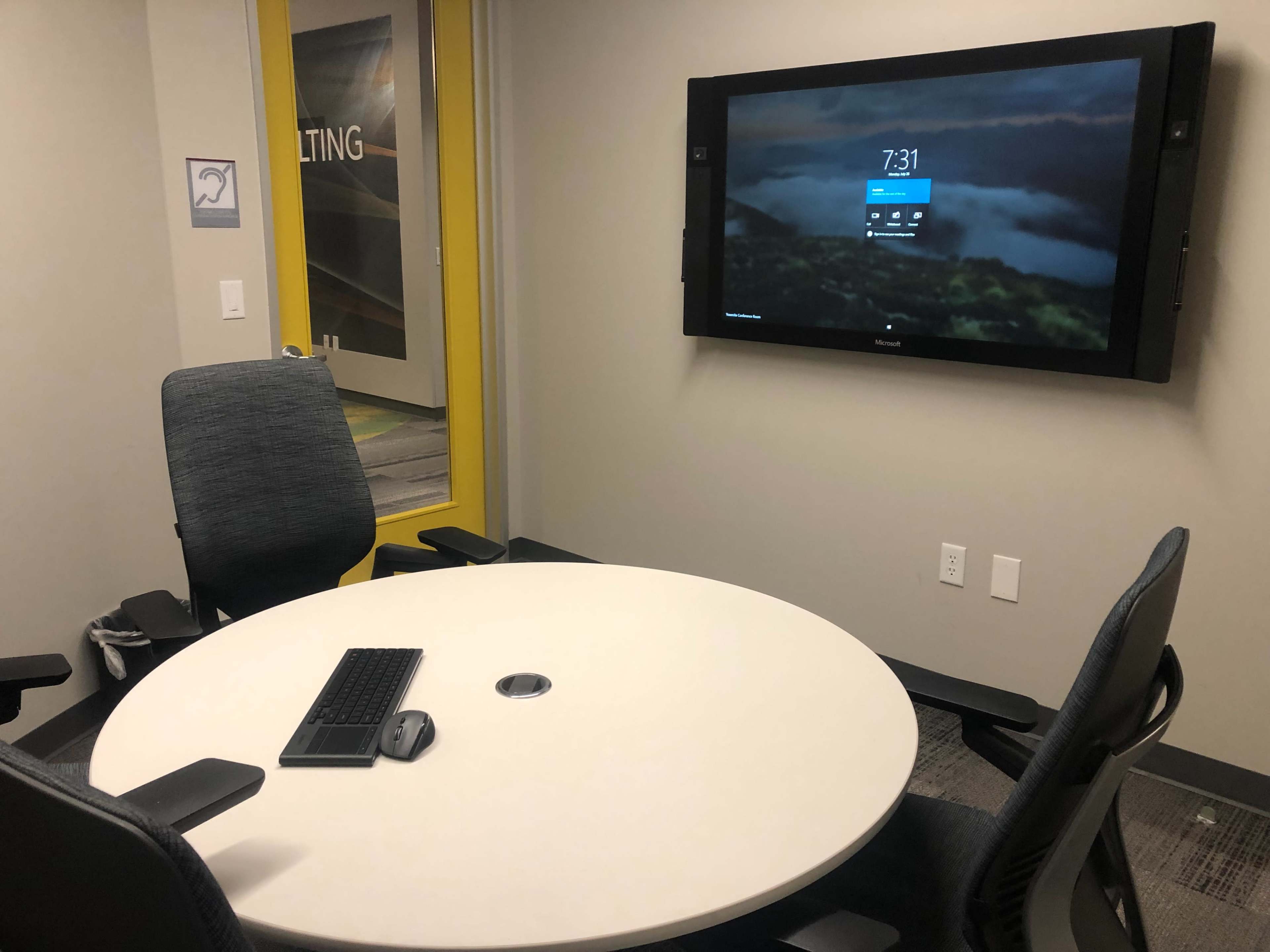 A conference room is set up with a circular table, four chairs, a computer keyboard, and a monitor displaying the time on the wall.