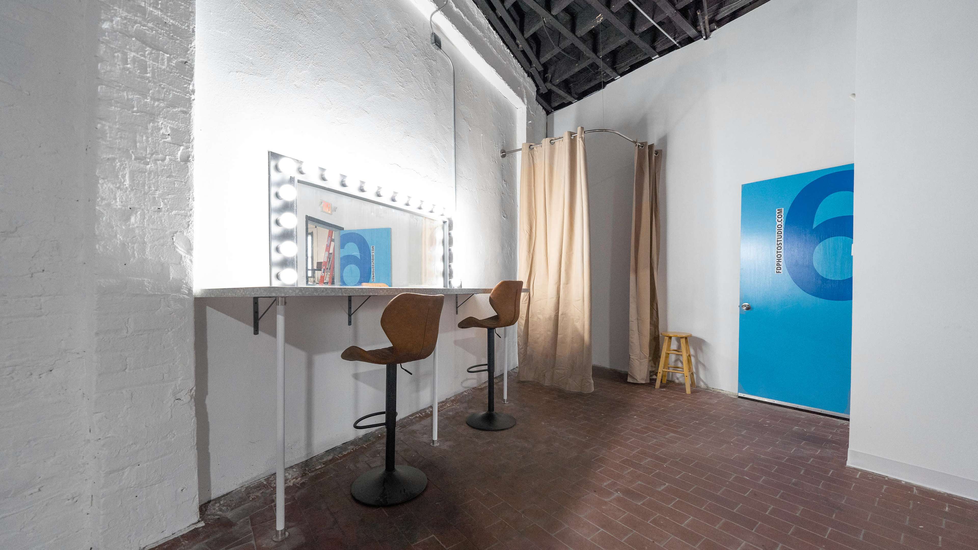 A minimalist dressing room with two high stools, a lighted mirror, and a blue door marked with the number 6.