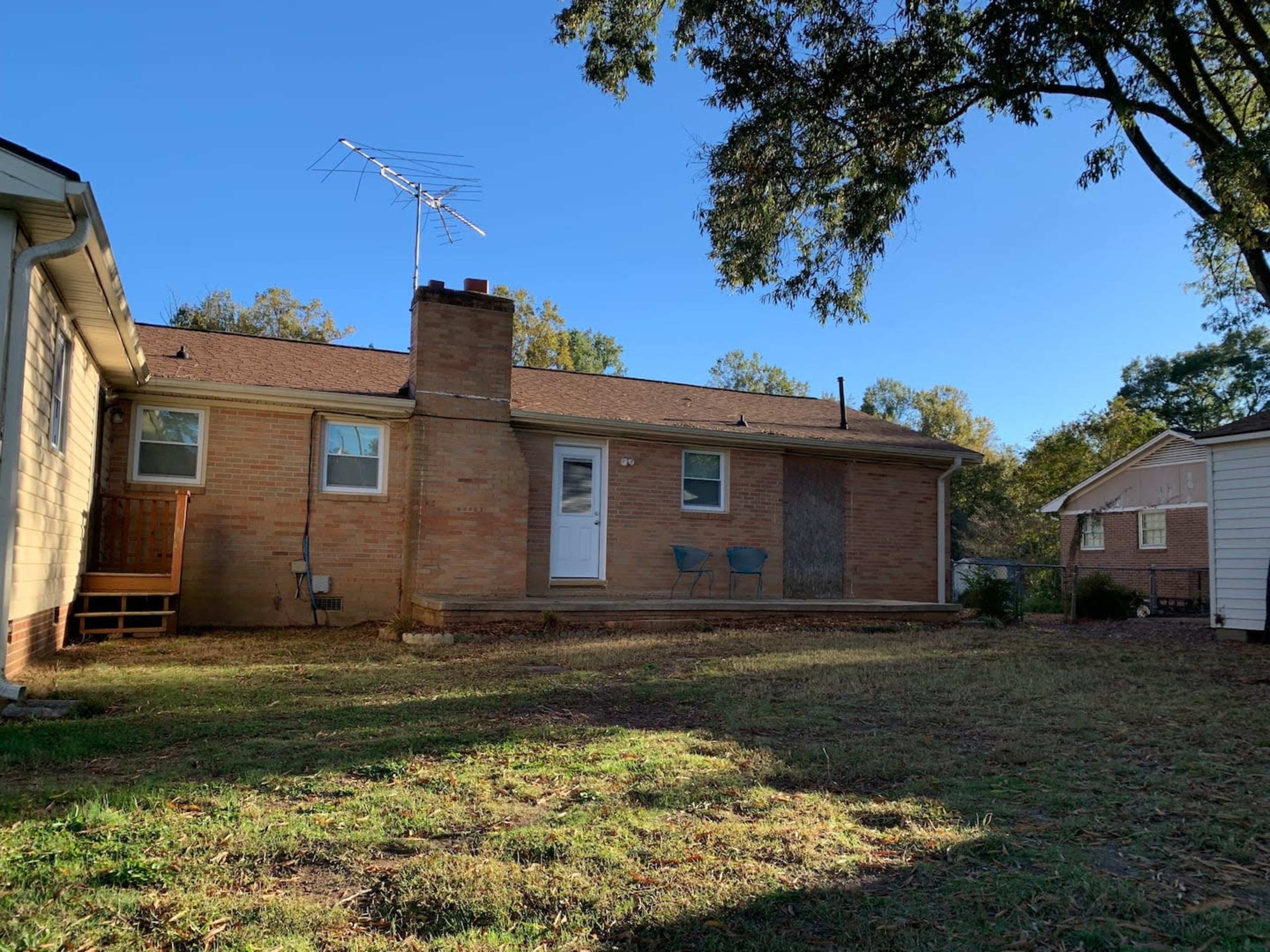 A brick house with a small porch, two blue chairs, and a chimney, surrounded by a grassy yard.