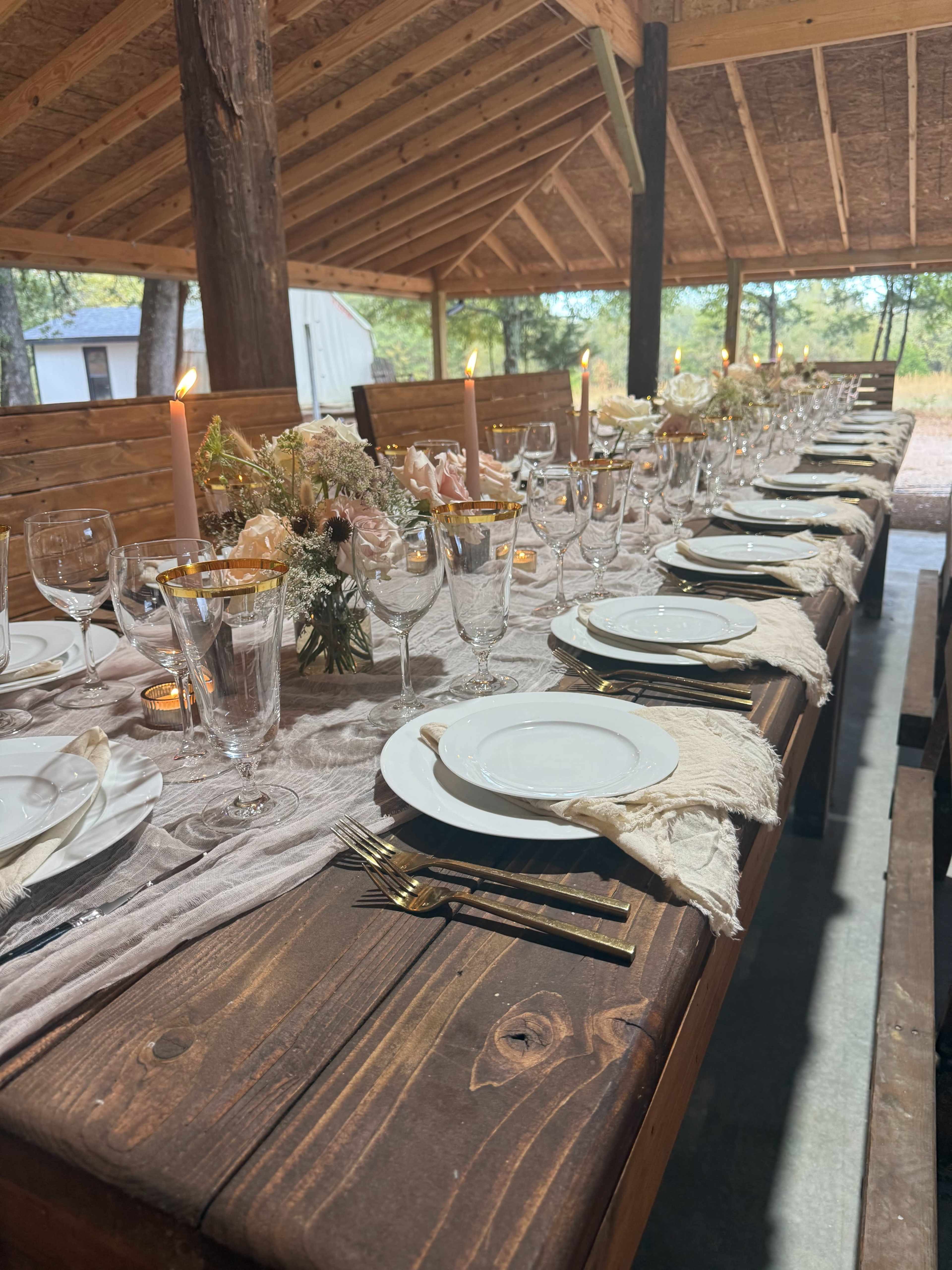 A long wooden table is set for a formal dinner outdoors, adorned with candles, floral arrangements, and elegant tableware.