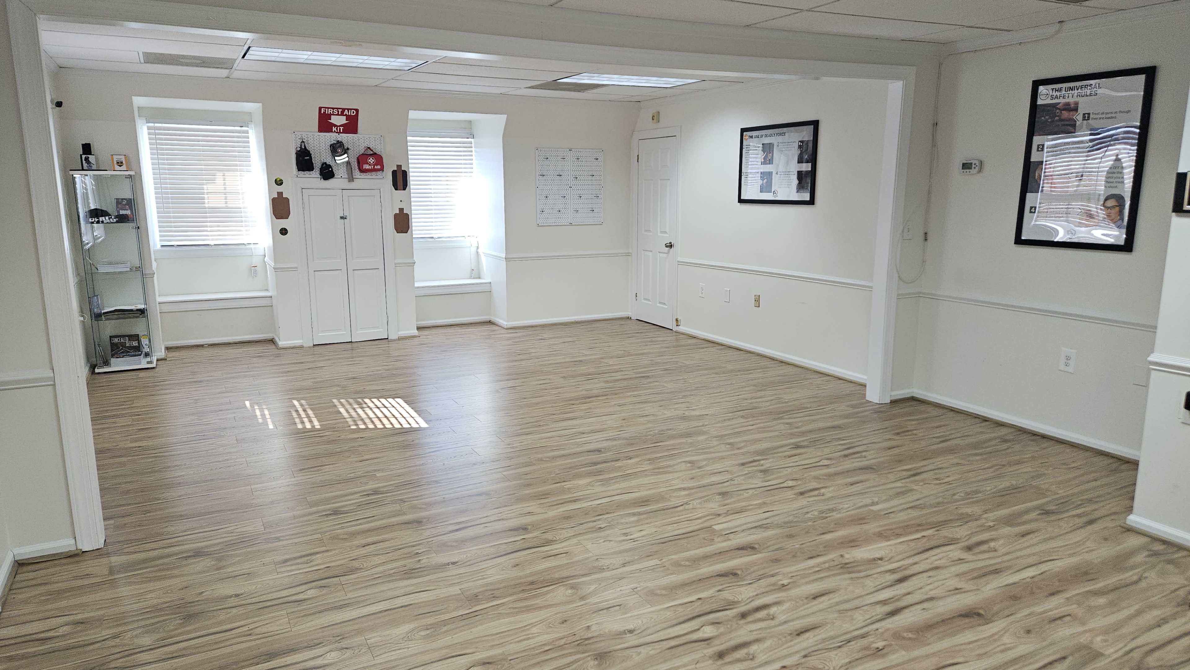 The image shows an empty room with wooden flooring, white walls, and two windows, along with a corner display case and a closed door.