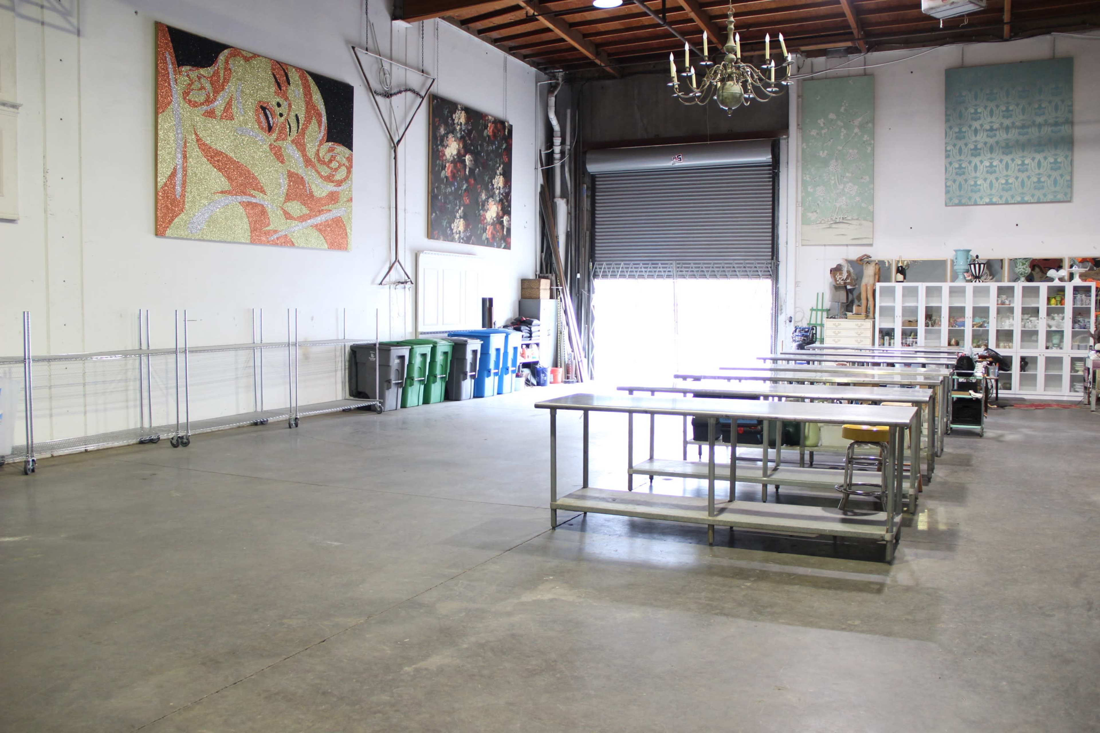 The image shows a spacious, unfinished interior with multiple work tables arranged in a row and large artworks hanging on the walls.