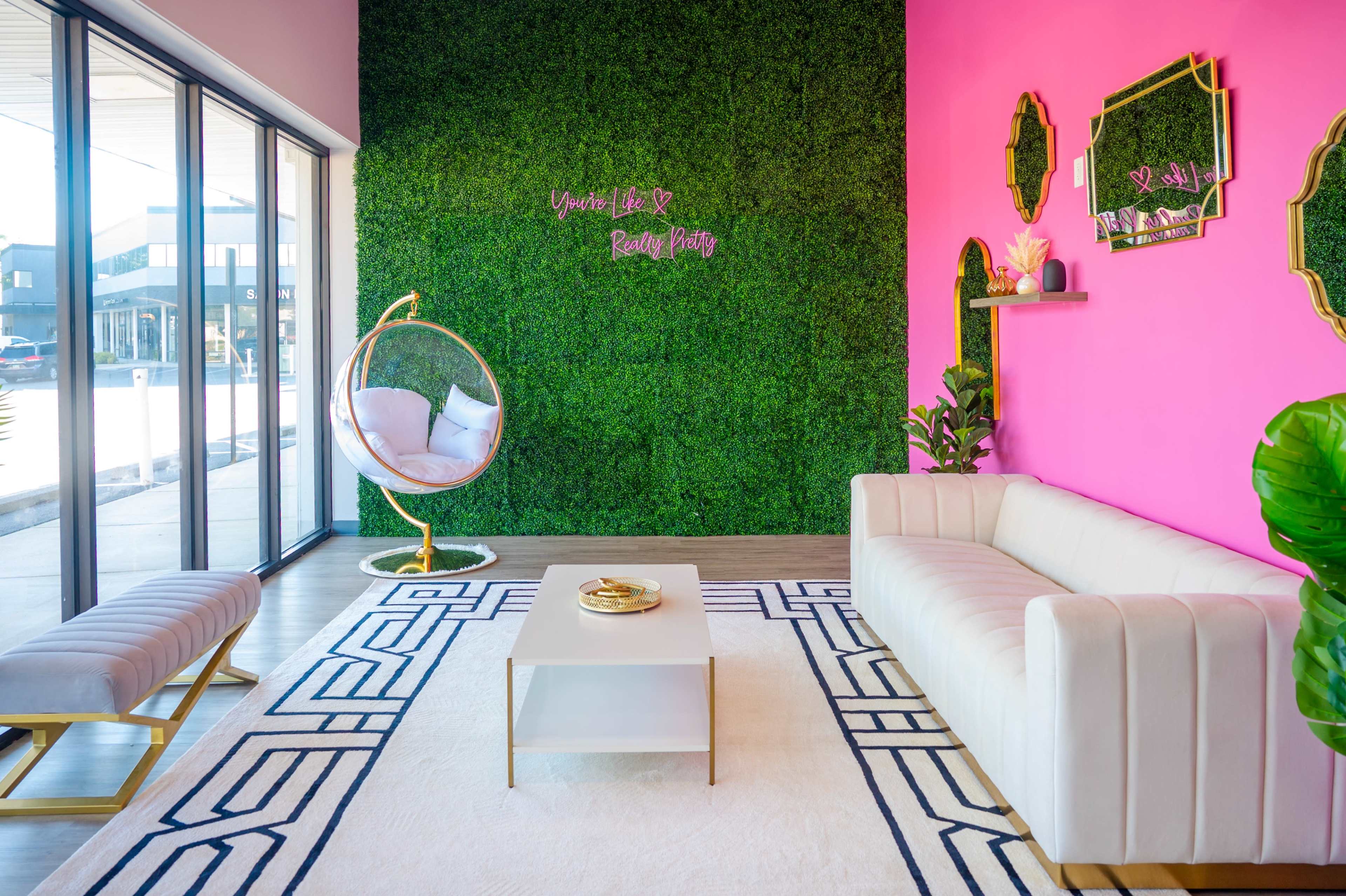 A bright interior space featuring a white sofa, a round hanging chair, and a green moss wall, all against a vibrant pink wall.