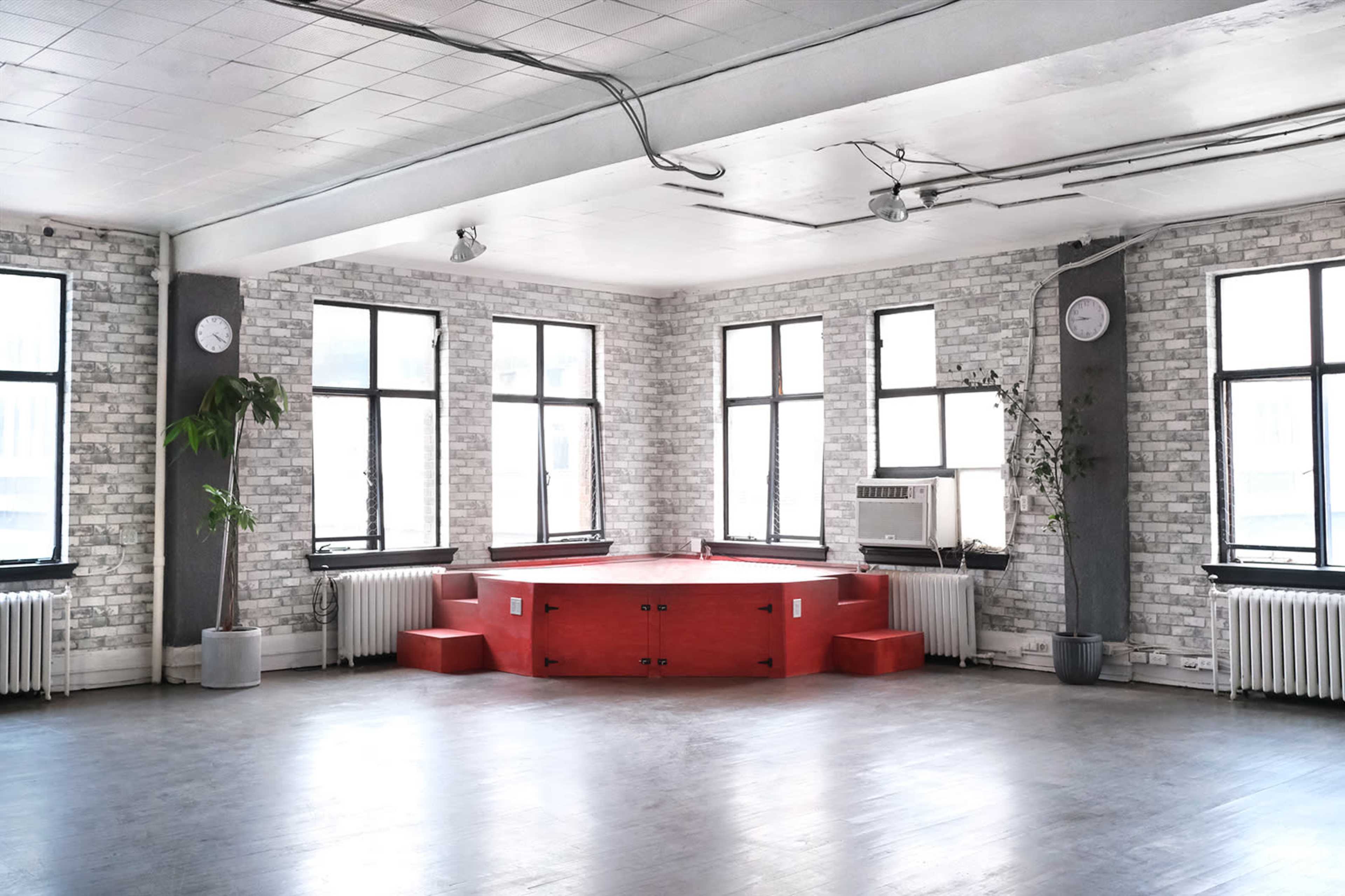 A spacious room with large windows, featuring a red platform in the center and industrial-style decor.