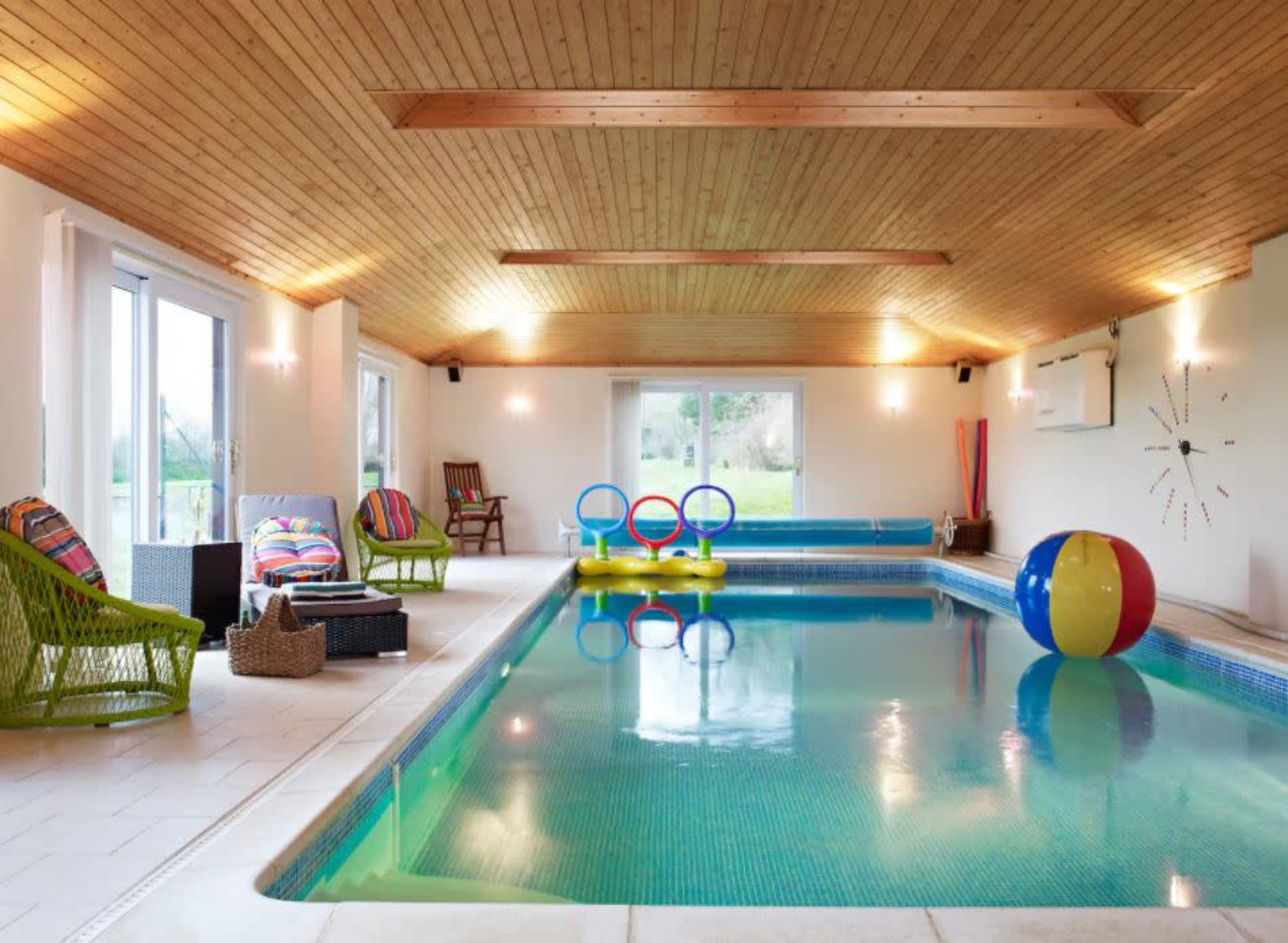 An indoor pool area featuring a rectangular swimming pool, colorful inflatable floats, and lounge chairs arranged around the space.