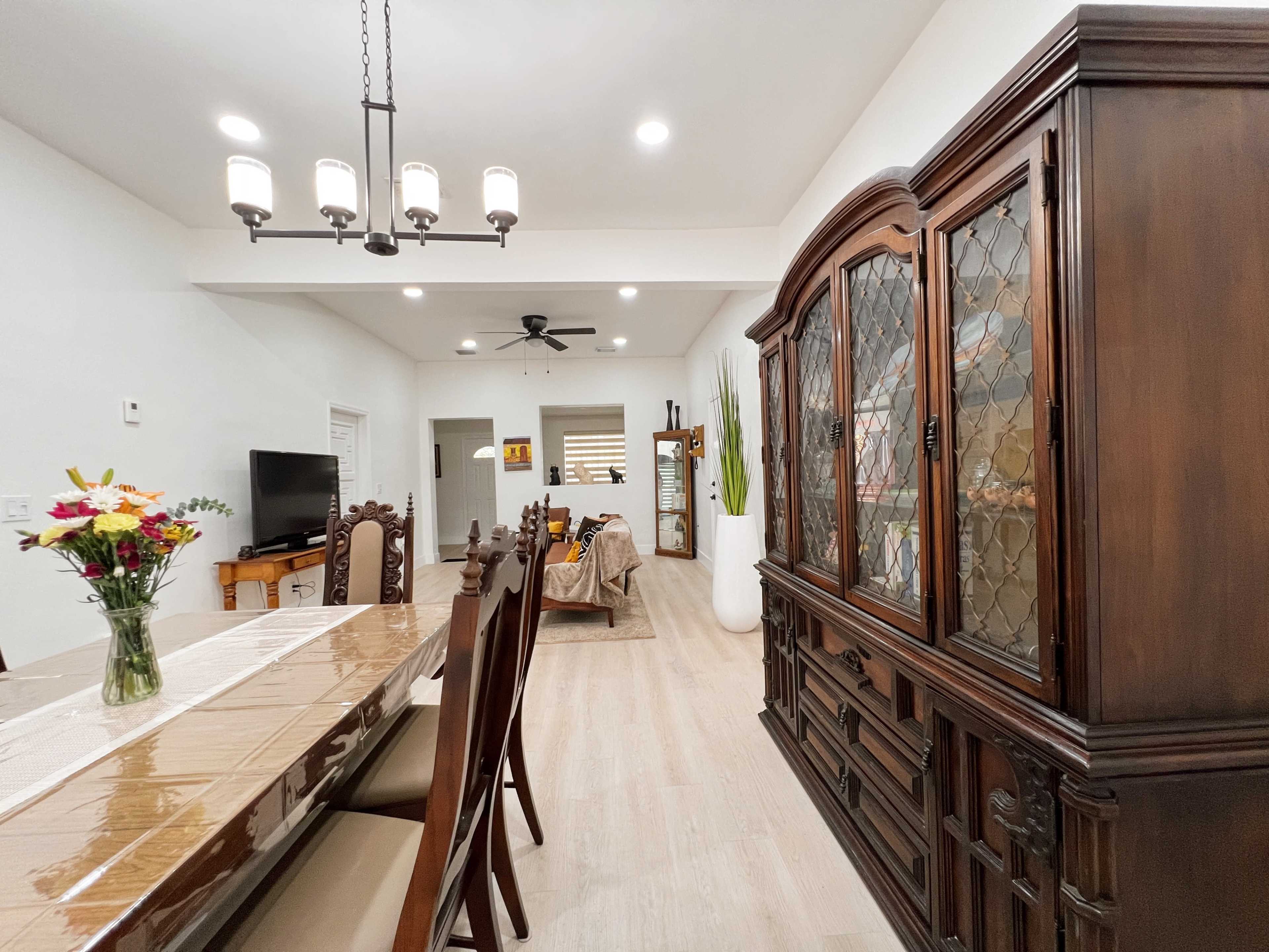 A dining area features a long table set with chairs, a wooden cabinet, and a living space visible in the background.