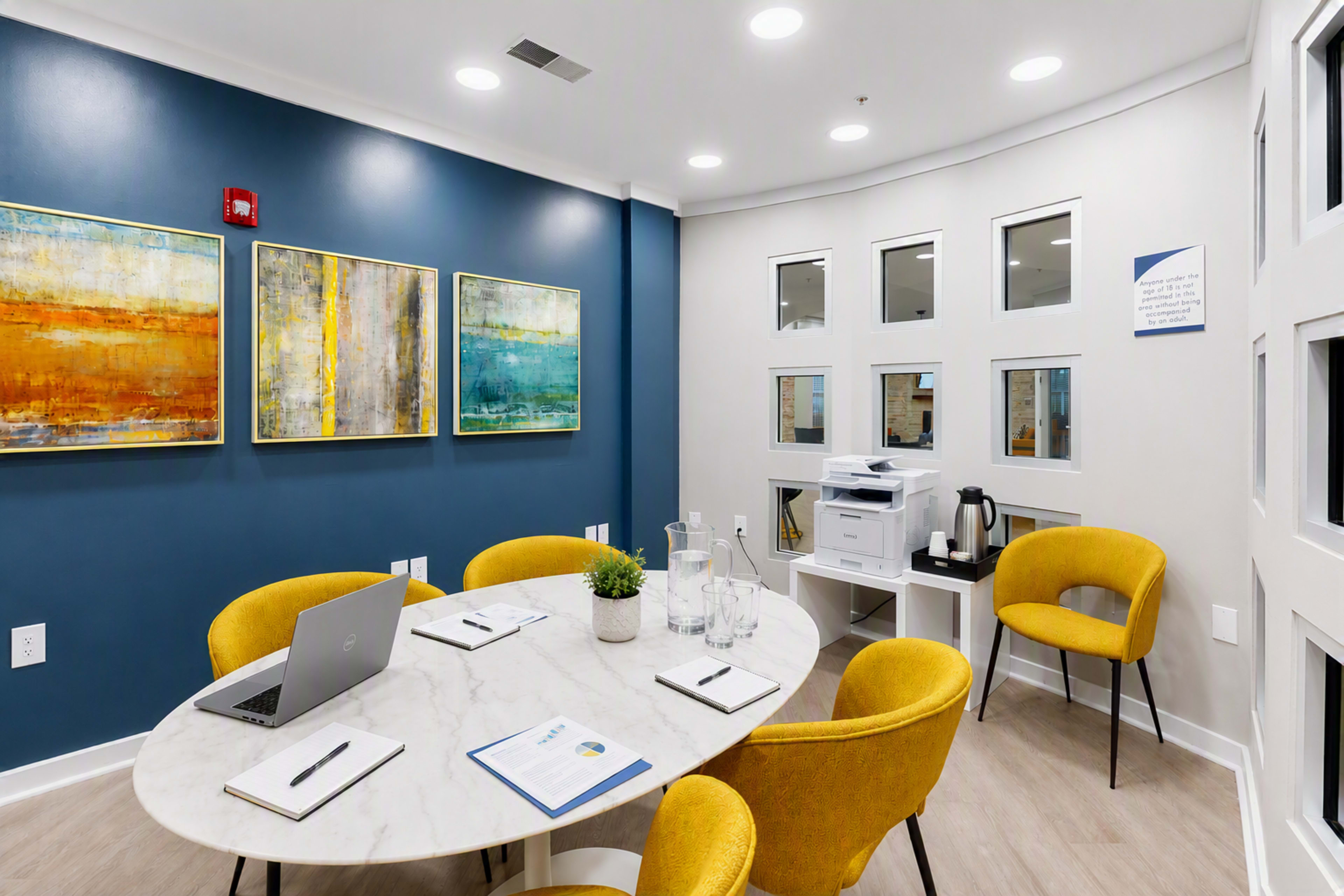 A modern conference room features a round marble table surrounded by yellow chairs, with framed artwork on the blue wall and a coffee station in the corner.