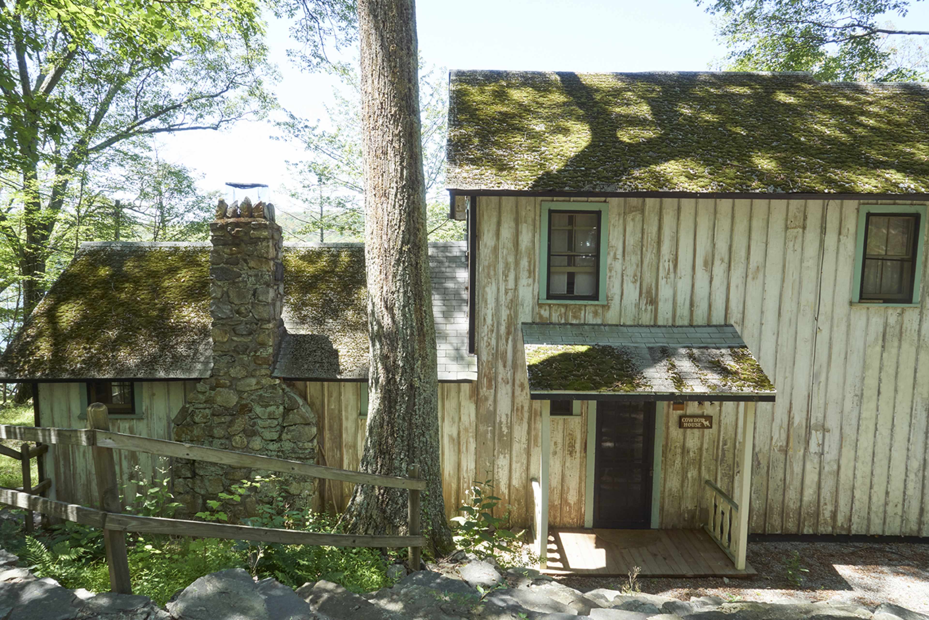 A two-story wooden cabin with a mossy roof and stone chimney is surrounded by trees and a grassy area.