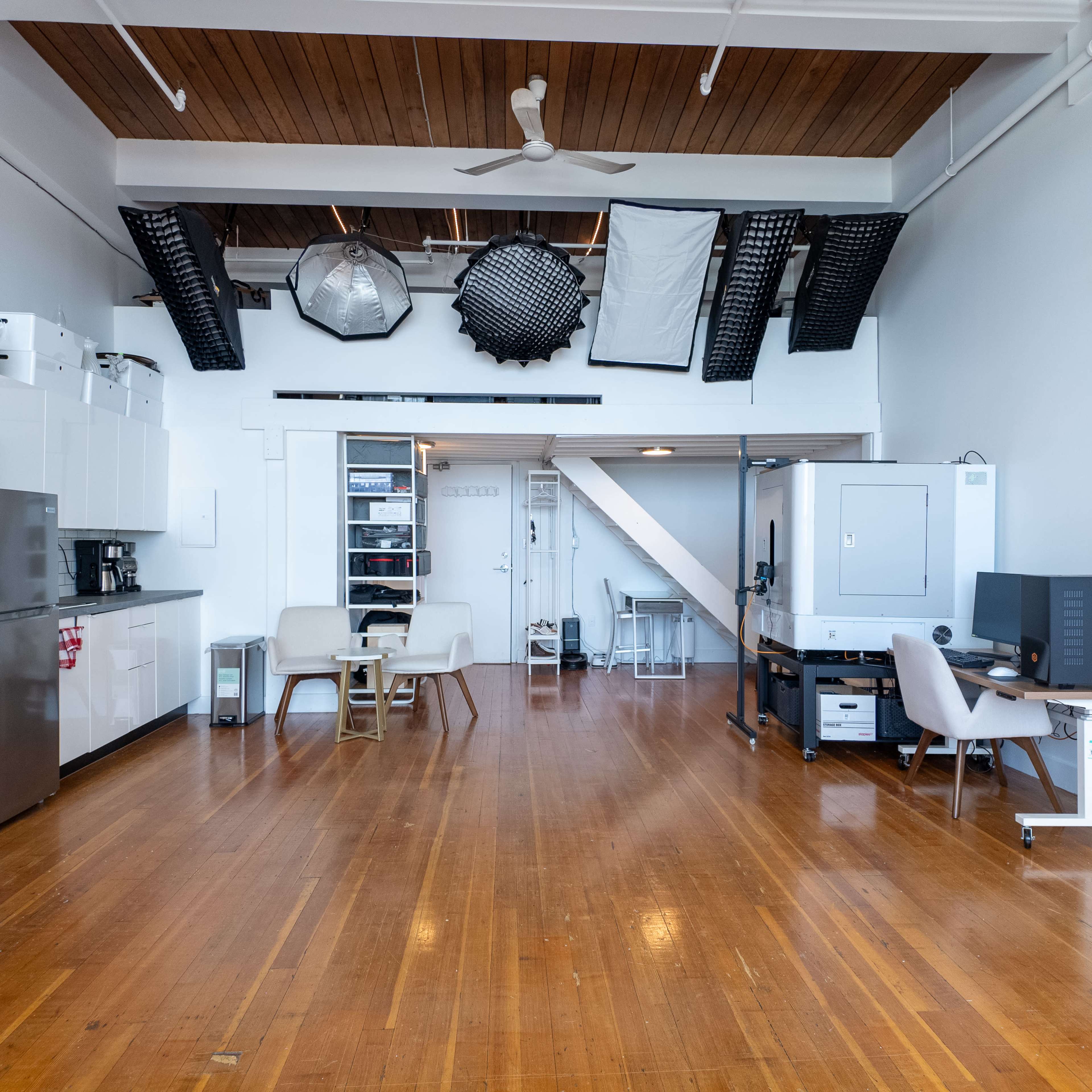 A bright, spacious studio with wooden flooring, equipped with kitchen cabinets, a dining table, and various lighting equipment hanging from the ceiling.