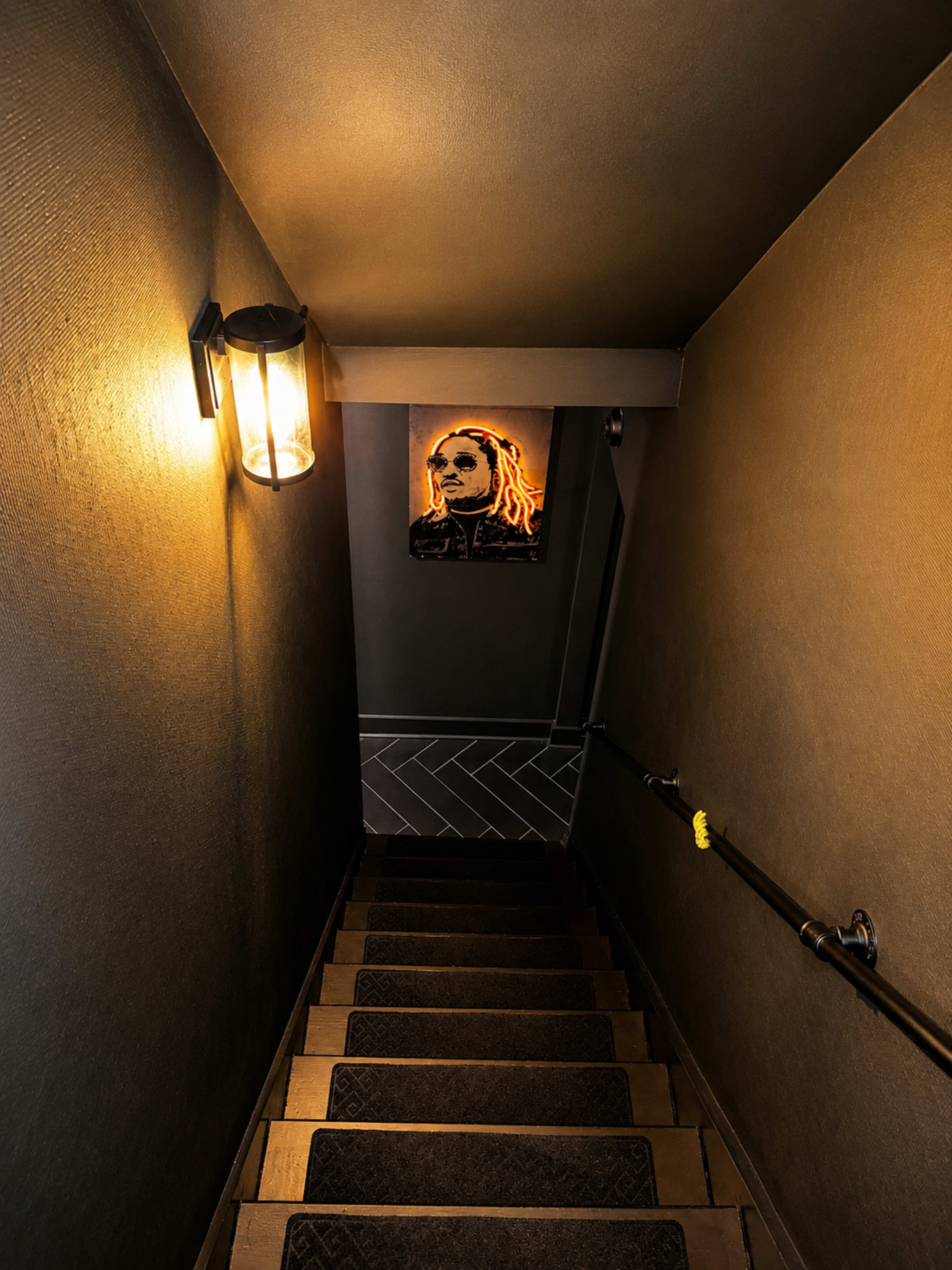 The image shows a dimly lit staircase with a handrail, leading downwards towards an illuminated wall art piece.