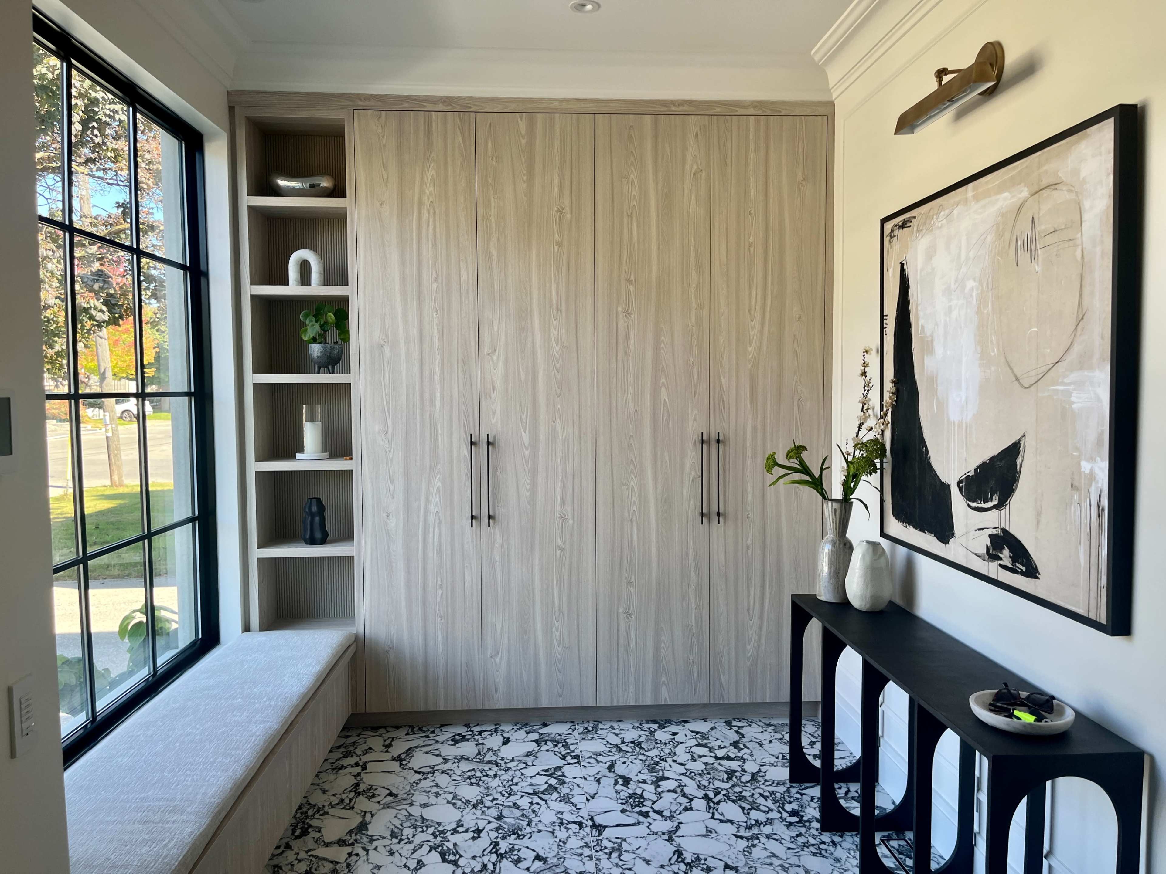 The image shows a modern entryway with wooden cabinetry, a marble-patterned floor, and a wall-mounted artwork, complemented by a bench and decorative items on shelves.