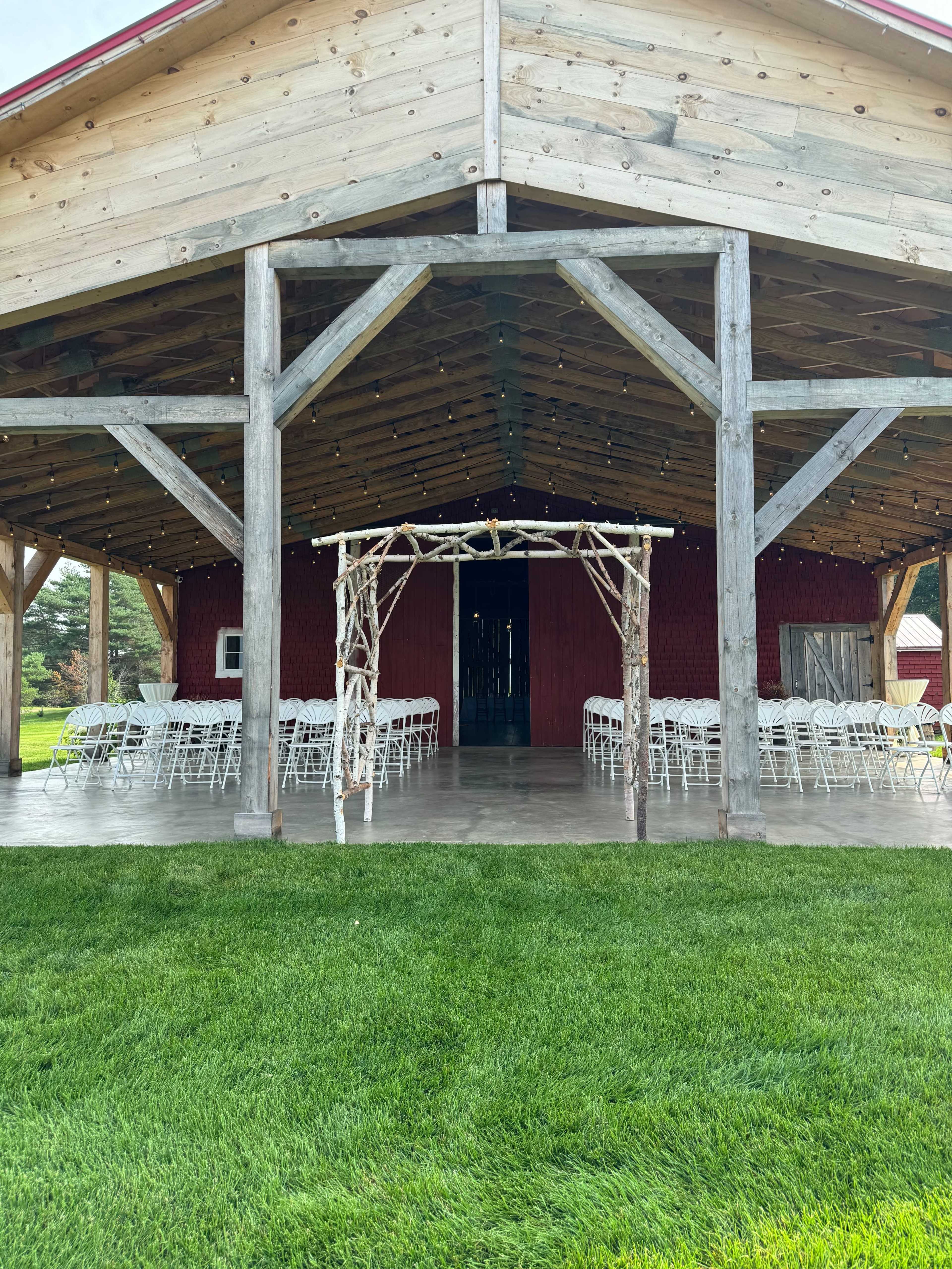 The image shows a rustic wooden barn with an open entrance, decorated with a wooden arch and set up for a gathering with white chairs arranged inside.