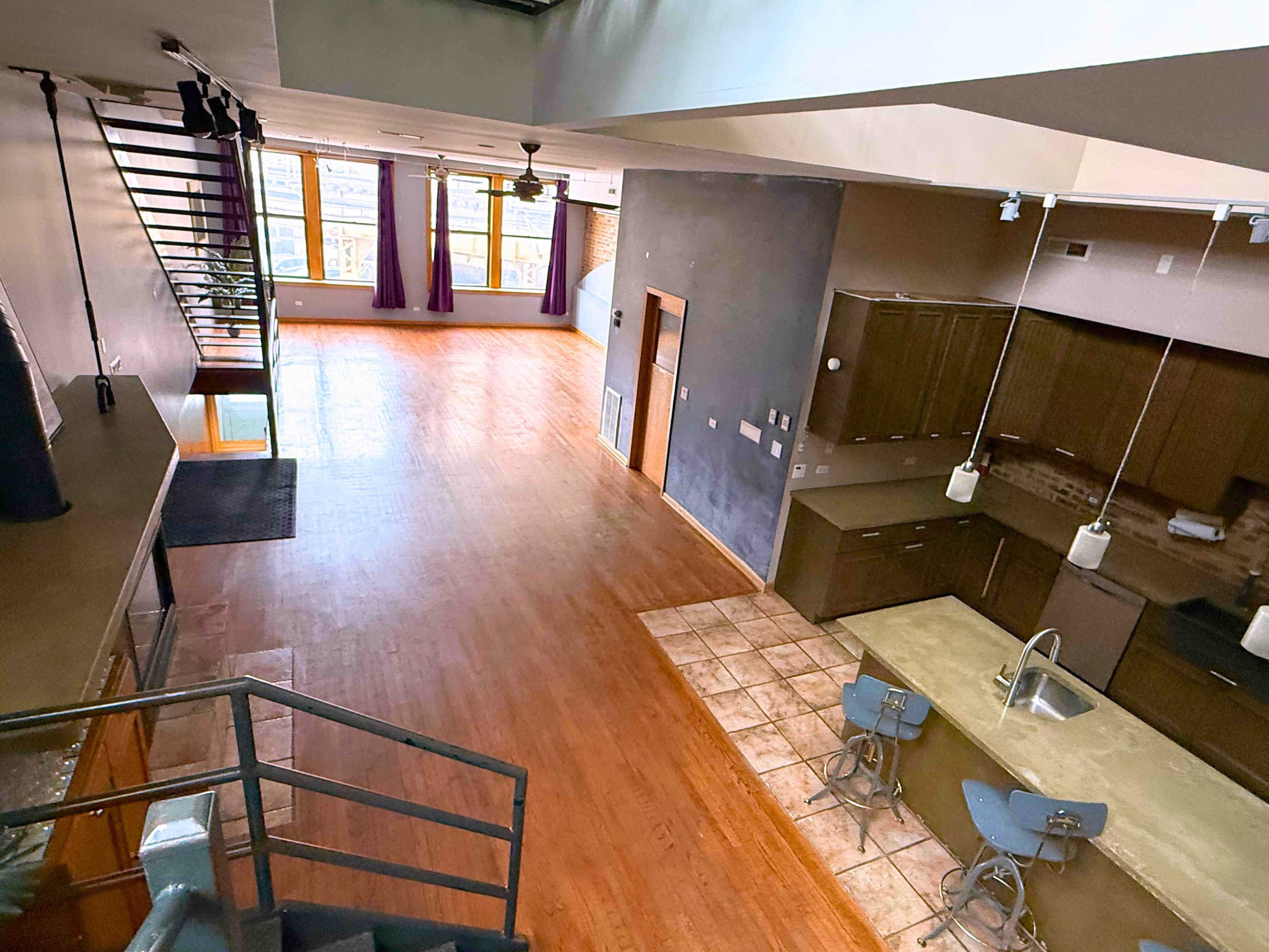 The image shows a spacious loft-style interior with wooden flooring, large windows, and an open kitchen area featuring modern appliances and a breakfast bar.