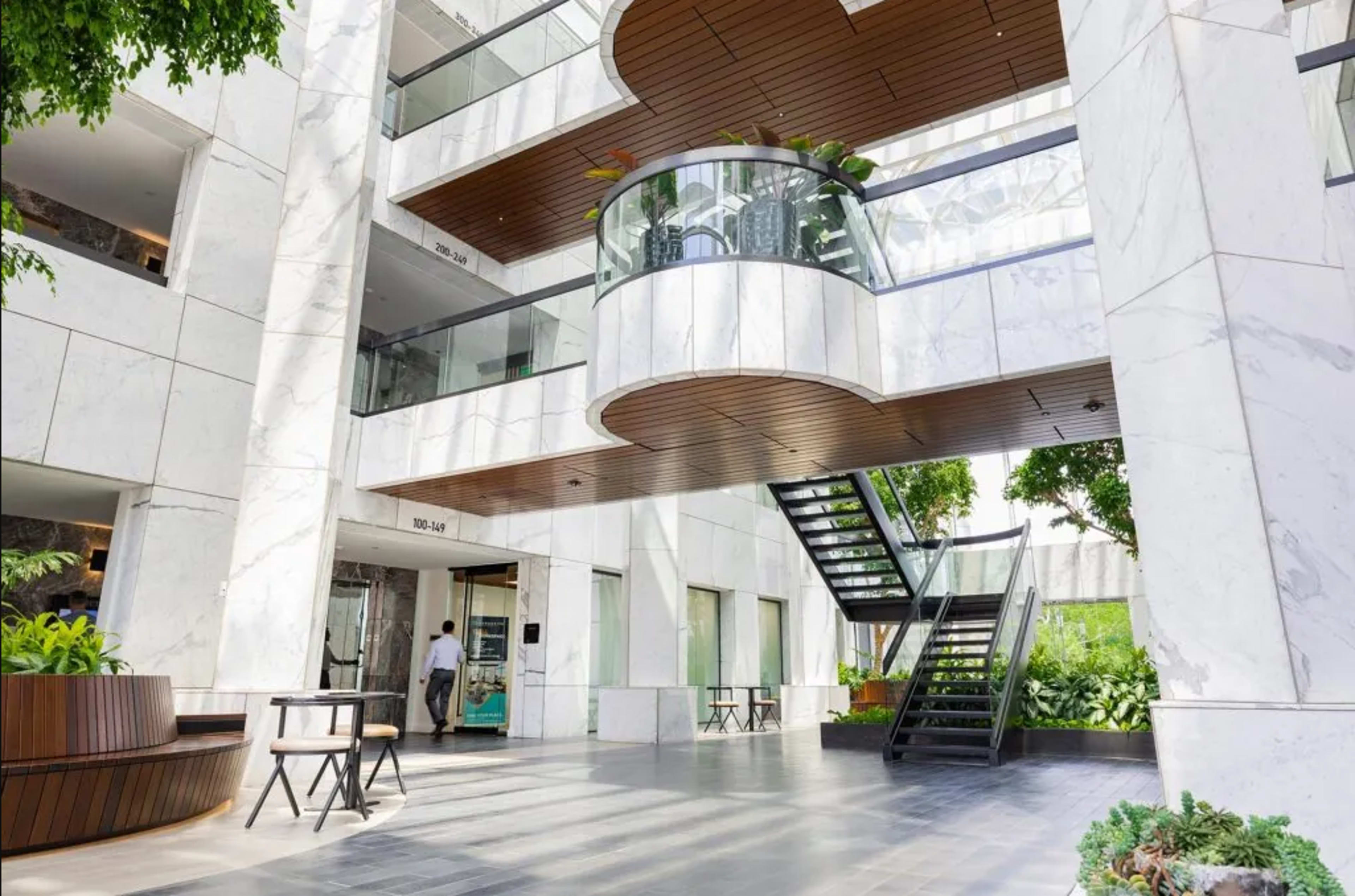 The image shows a modern indoor atrium featuring architectural marble finishes, glass railings, and a central staircase surrounded by greenery.