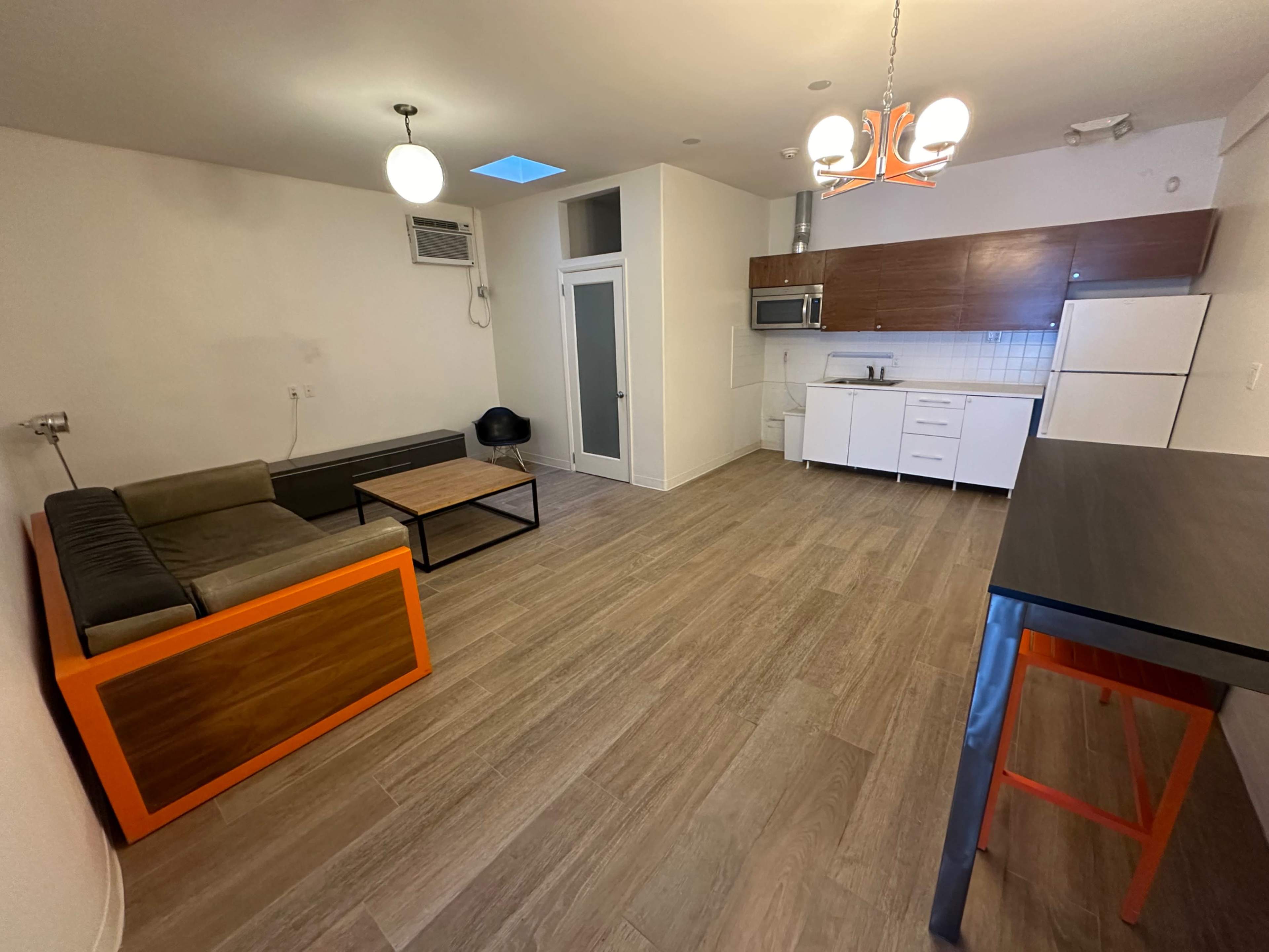 The image shows a modern, minimalist apartment interior featuring a sofa, coffee table, kitchen area, and dining bar.