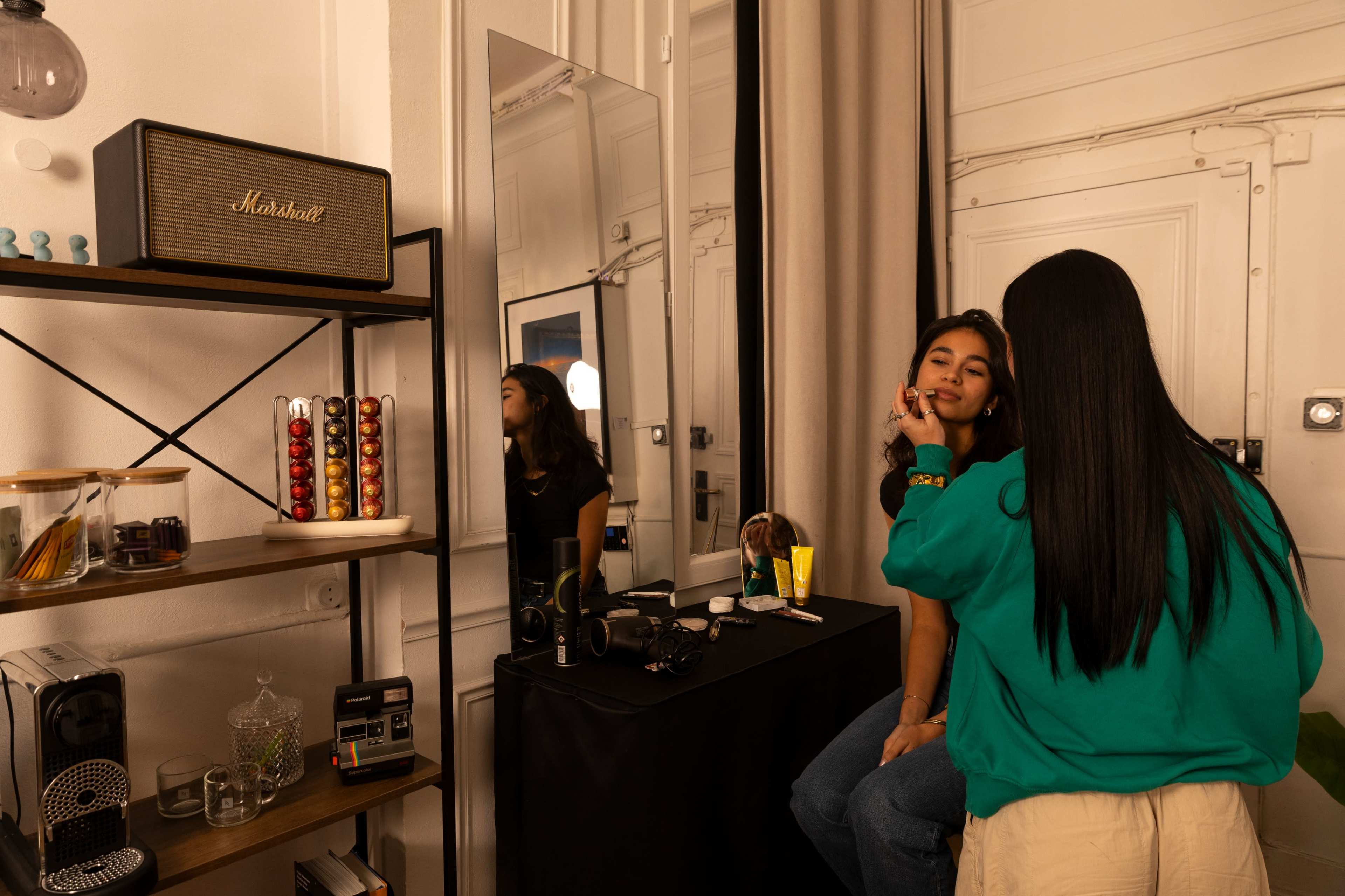 A person is applying makeup to another person in a brightly lit room with a mirror and a vintage speaker on a shelf.