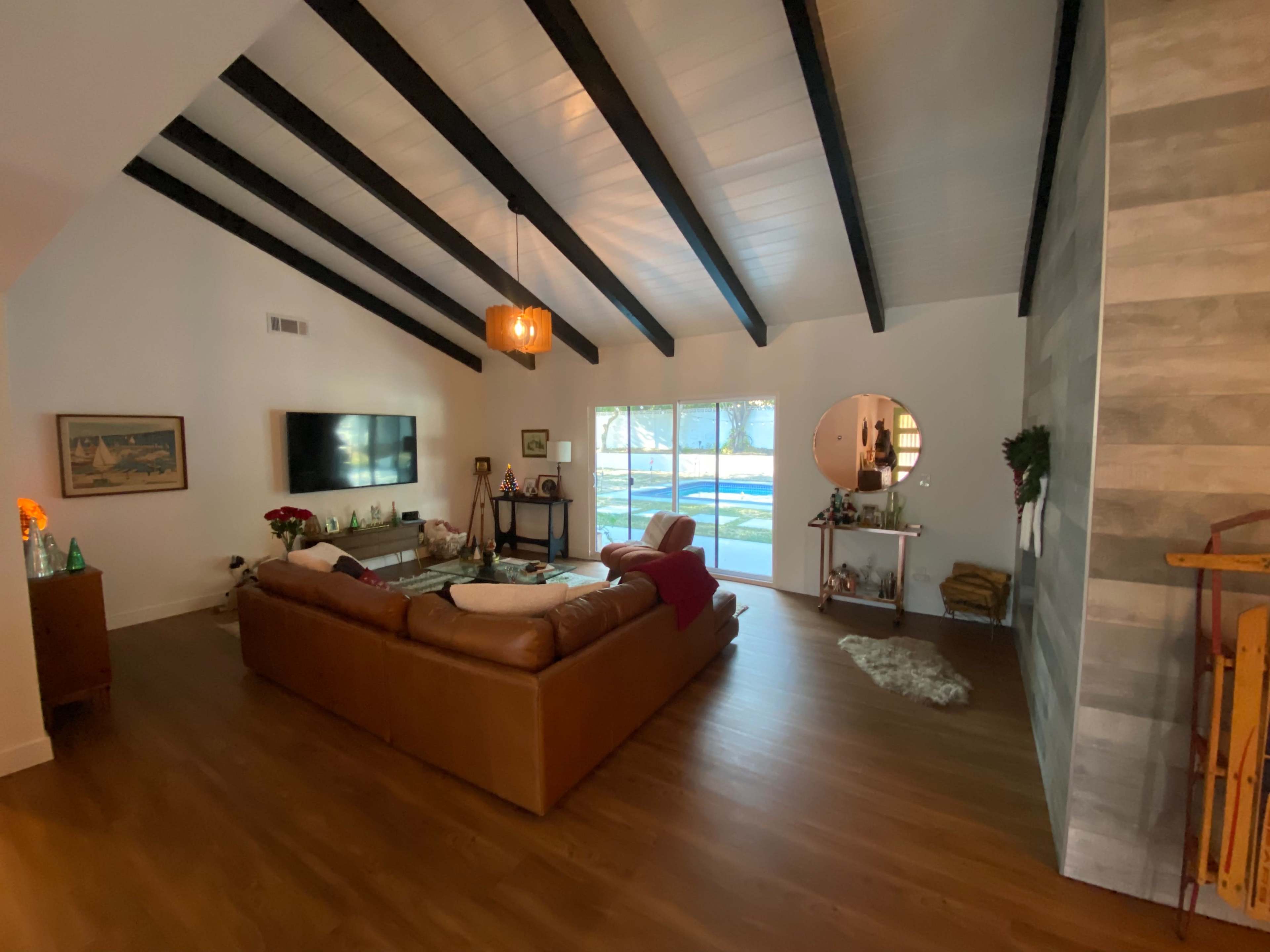 The image shows a living room with a high vaulted ceiling, featuring a brown leather sofa, large windows allowing natural light, and a small decorative table.
