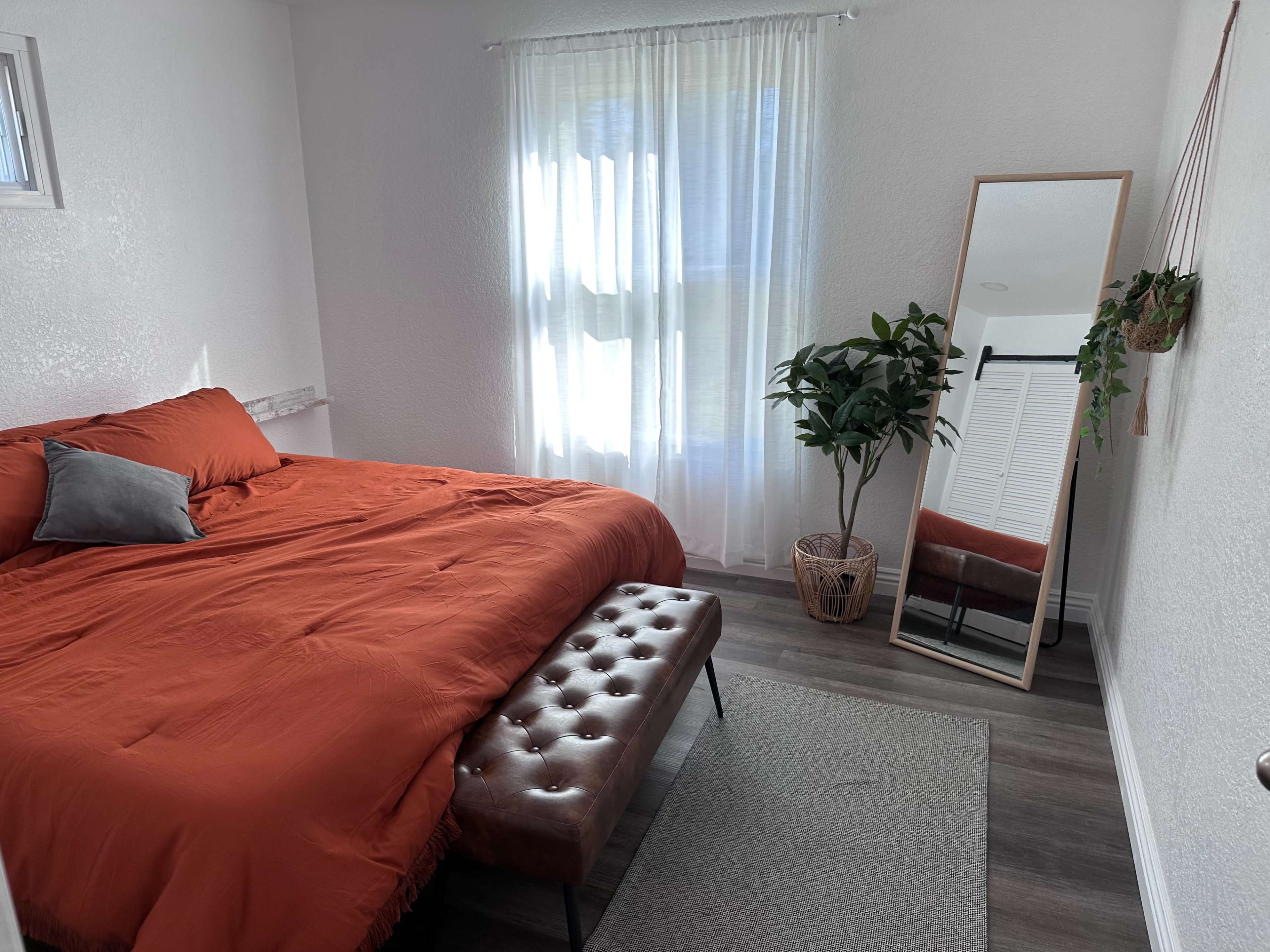 A bright bedroom featuring an orange bedspread, a mirror, a potted plant, and a bench at the foot of the bed.