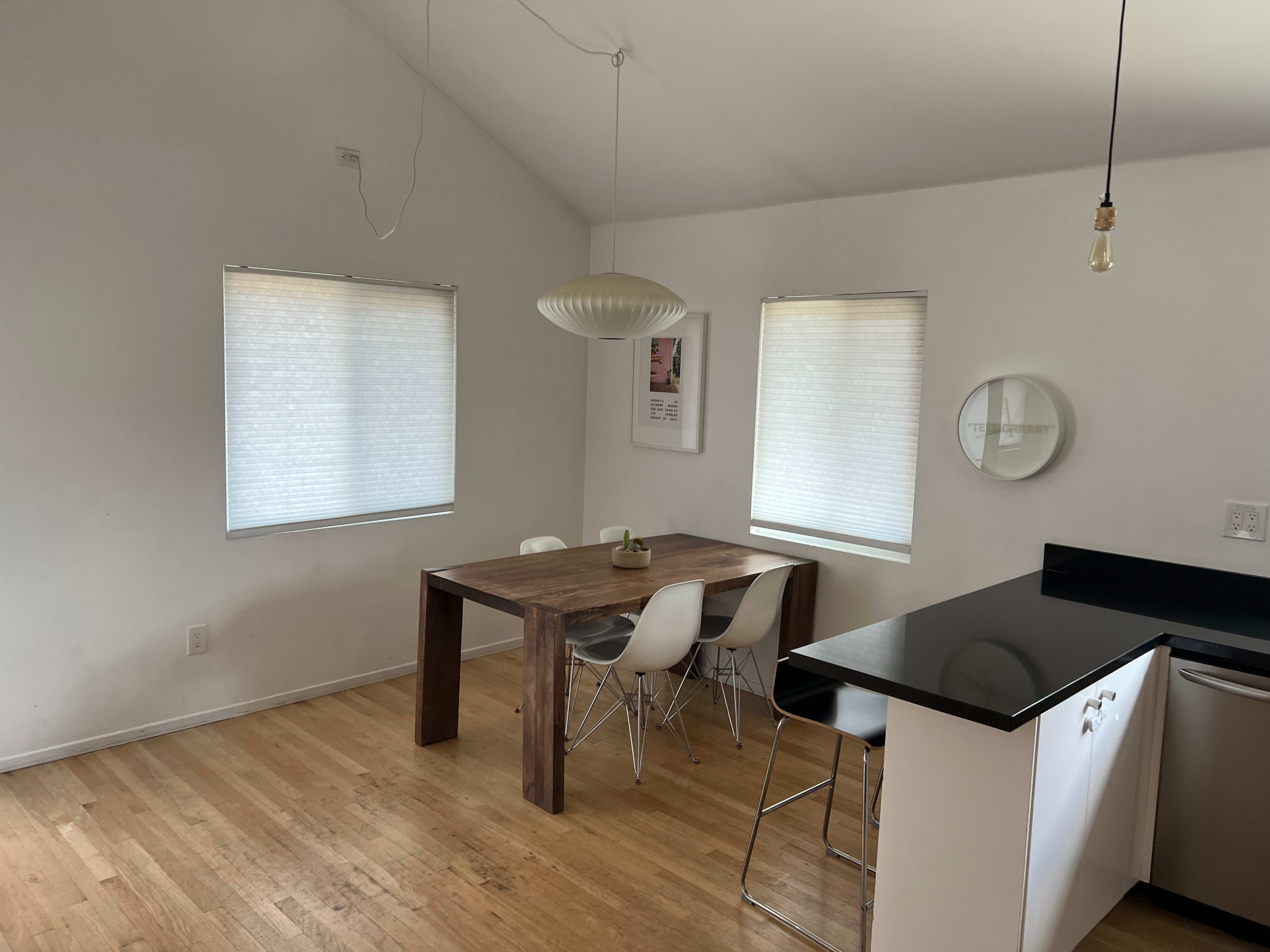 The image depicts a minimalist dining area featuring a wooden table with white chairs, a pendant light, and large windows with blinds.