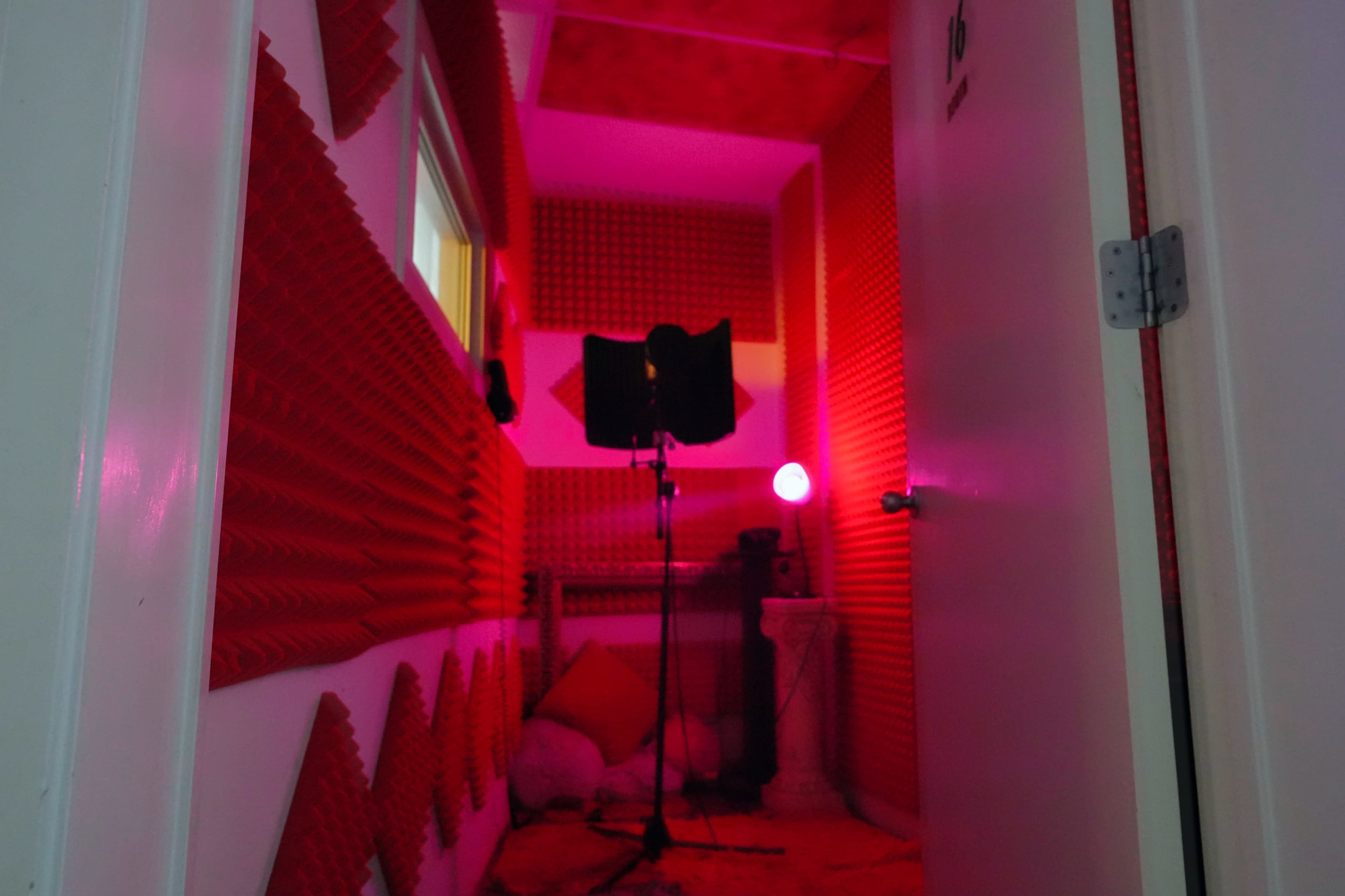 The image shows a recording booth lined with red soundproofing panels, featuring a microphone stand and a glowing lamp.