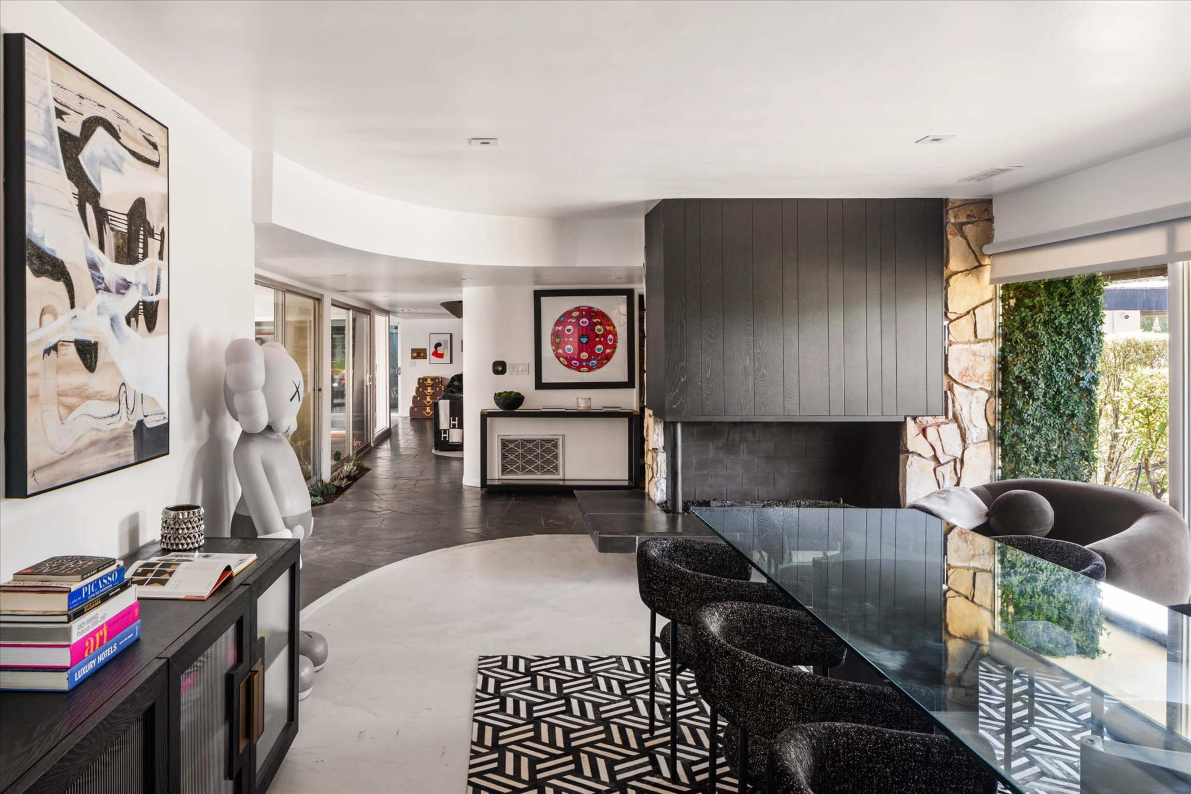 A modern dining area features a glass table surrounded by black chairs, with an artwork-adorned wall and a stone fireplace in the background.
