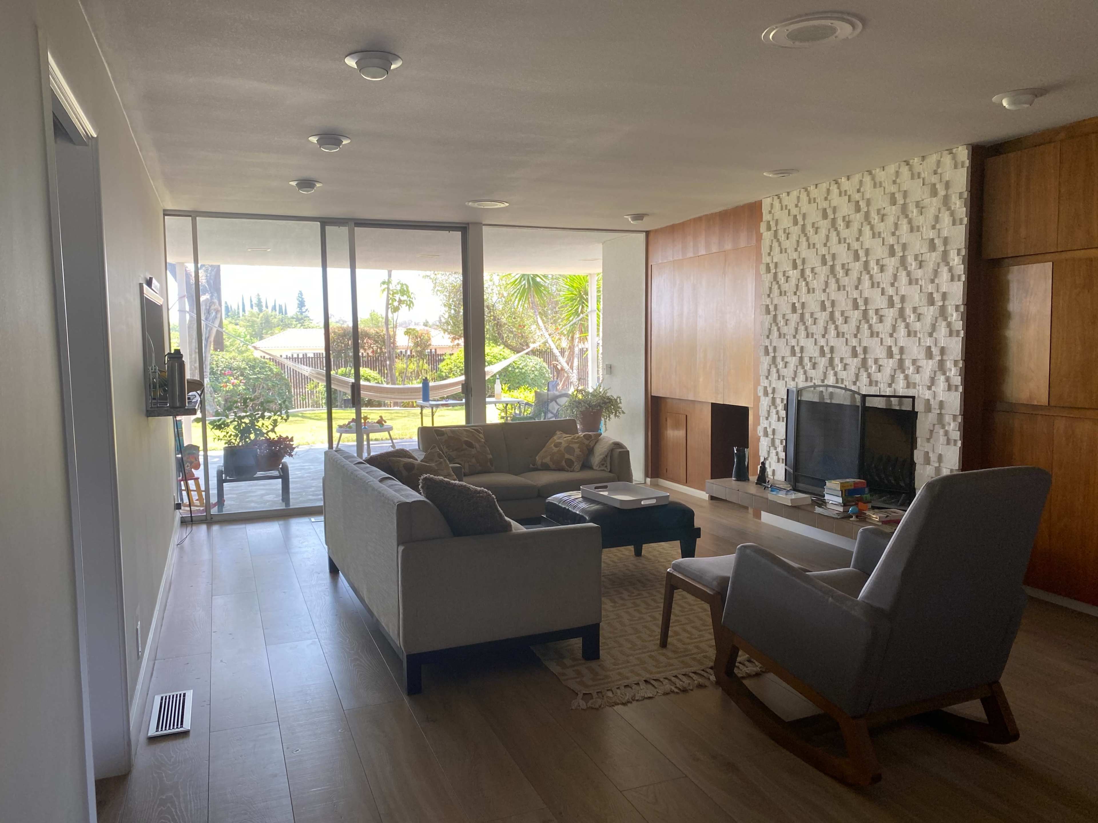 A living room features a sofa, an armchair, and a fireplace, with large windows overlooking a backyard garden.