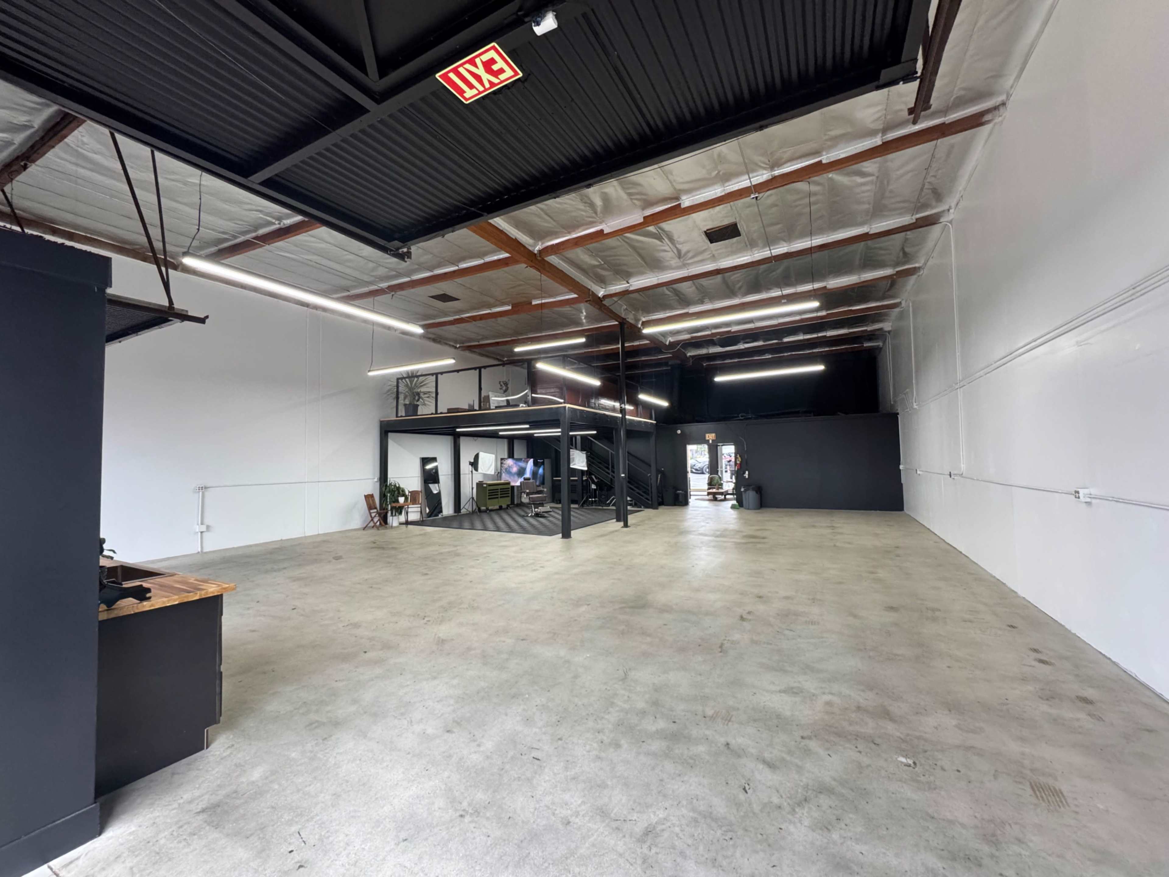 The image shows a spacious, empty industrial-style room with exposed beams, polished concrete flooring, and a staircase in the background.