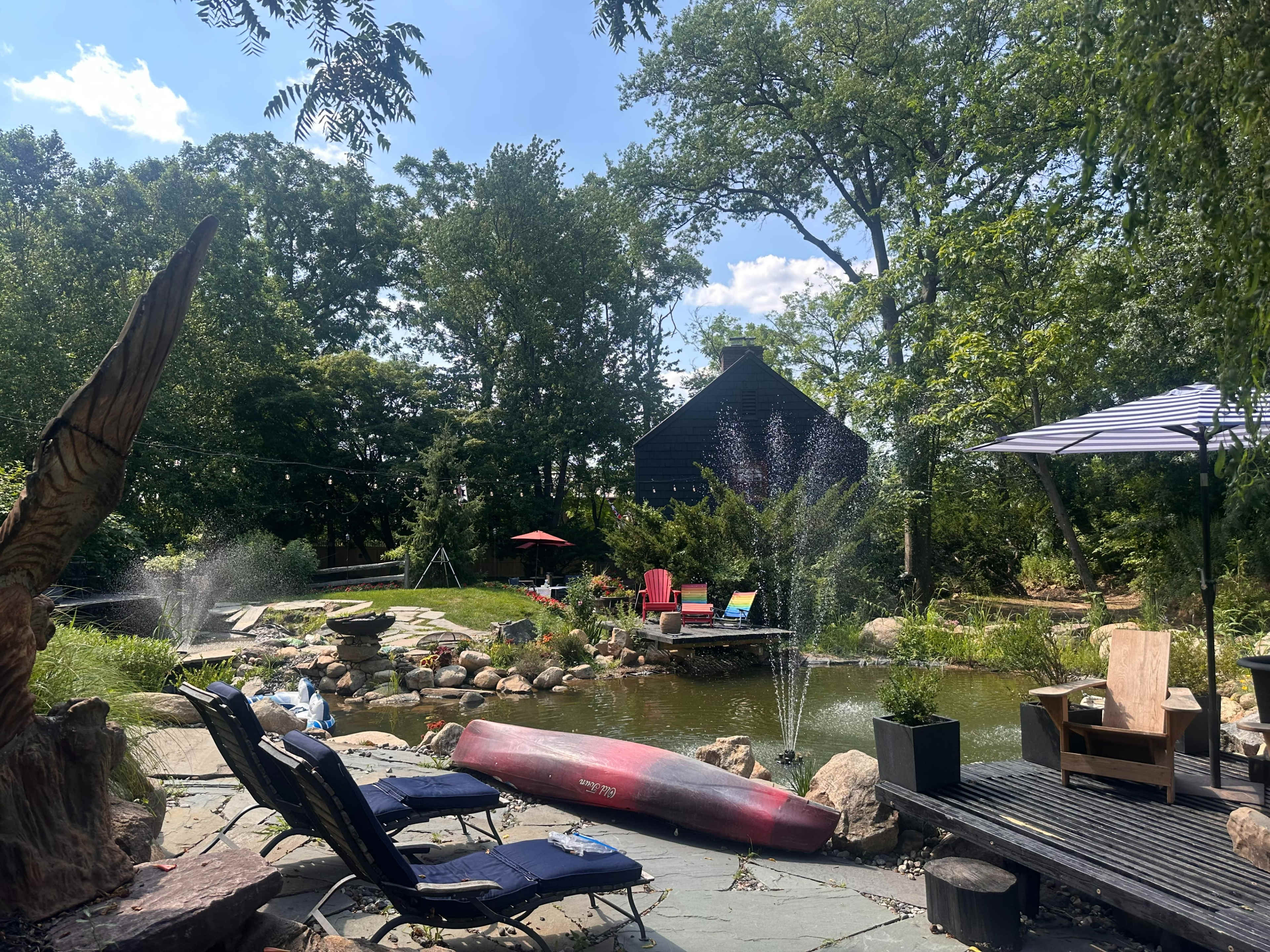 The image features a serene outdoor space with a pond surrounded by rocks, lounge chairs, and a kayak, with a house and trees in the background.