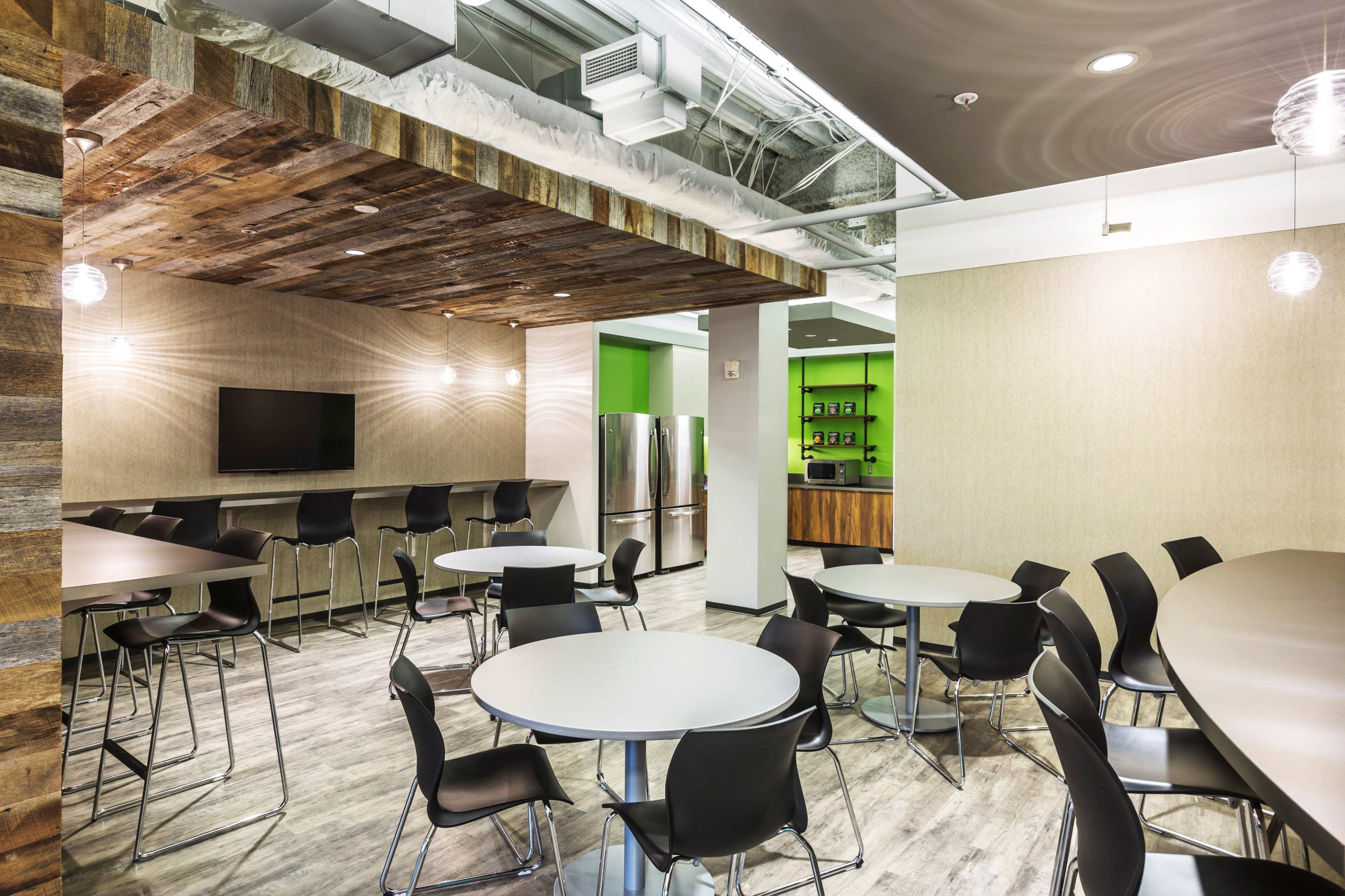 A modern communal kitchen area with tables, chairs, a television, and a kitchen counter featuring an open layout and wooden accents.