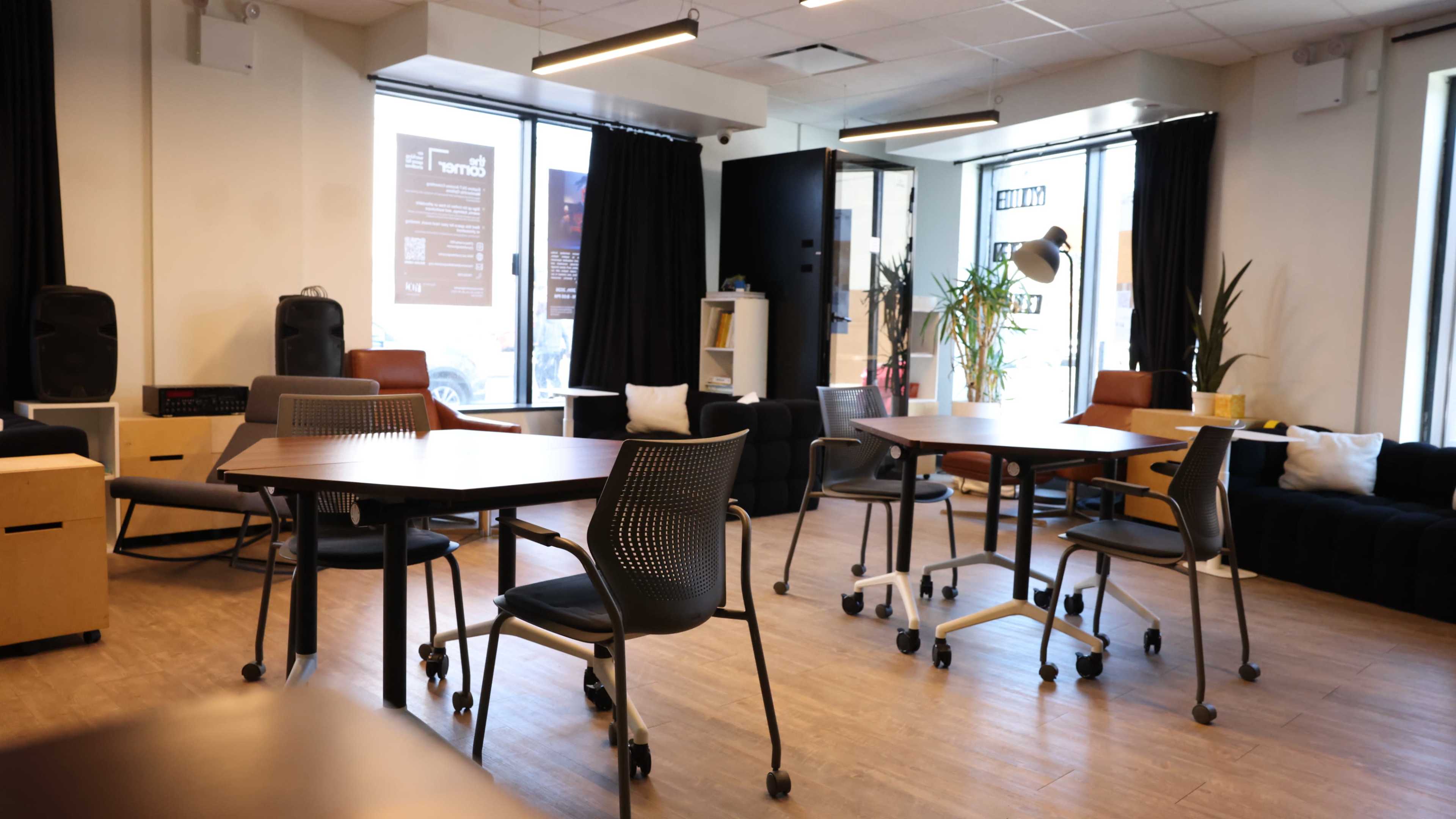 The image shows a modern, open workspace with several tables and chairs arranged around the room, featuring large windows letting in natural light.
