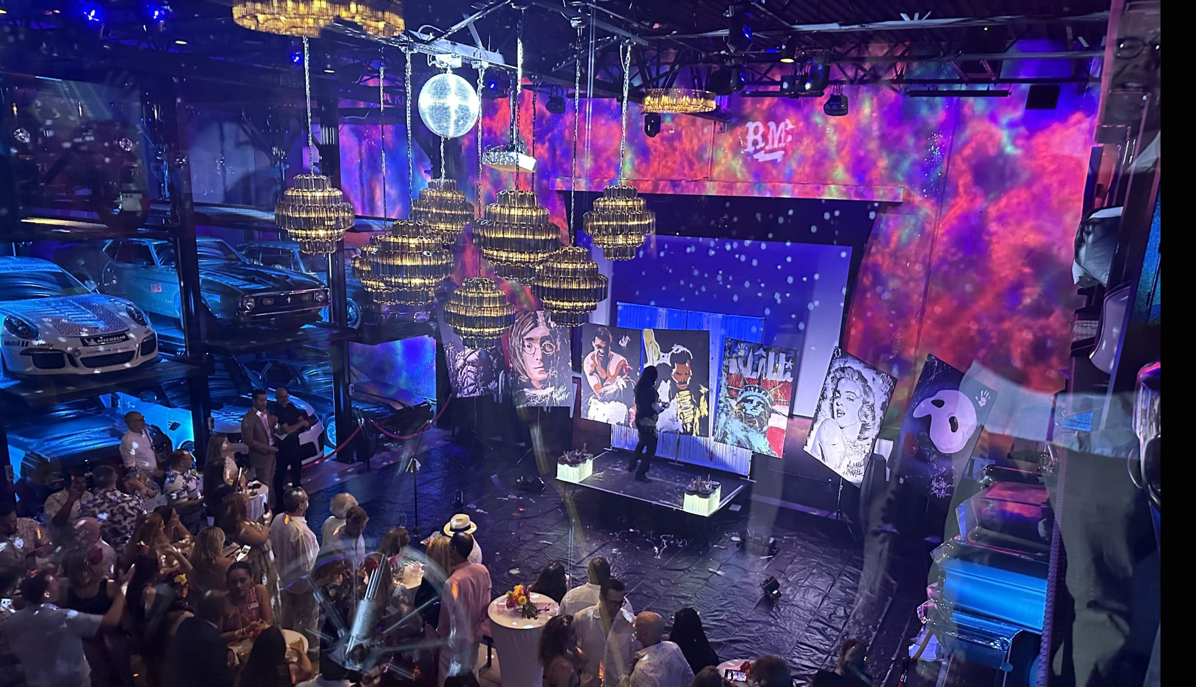 A vibrant indoor event space features a stage with a performer, surrounded by art displays and suspended lights, while guests mingle below amidst colorful projections and vintage cars.