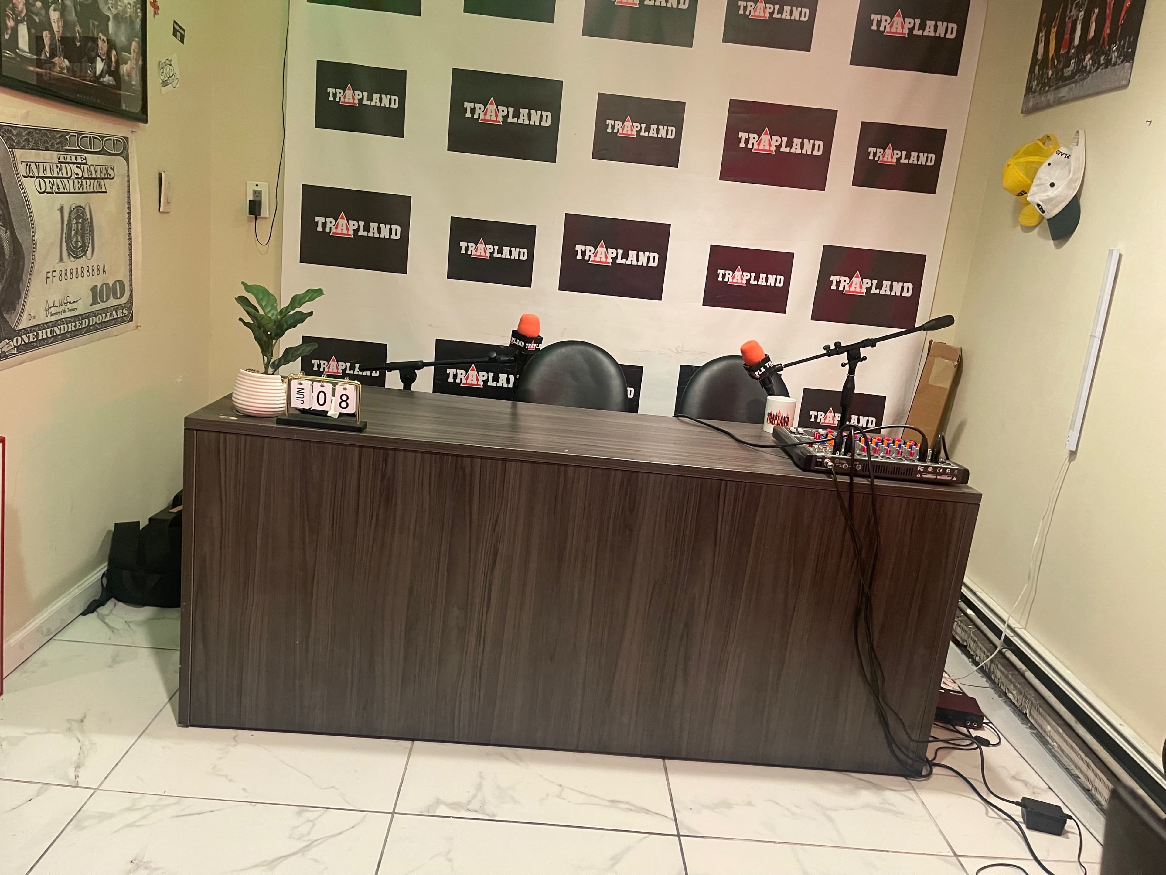 A recording studio setup with a wooden table, two chairs, and microphones, against a backdrop of branded posters.