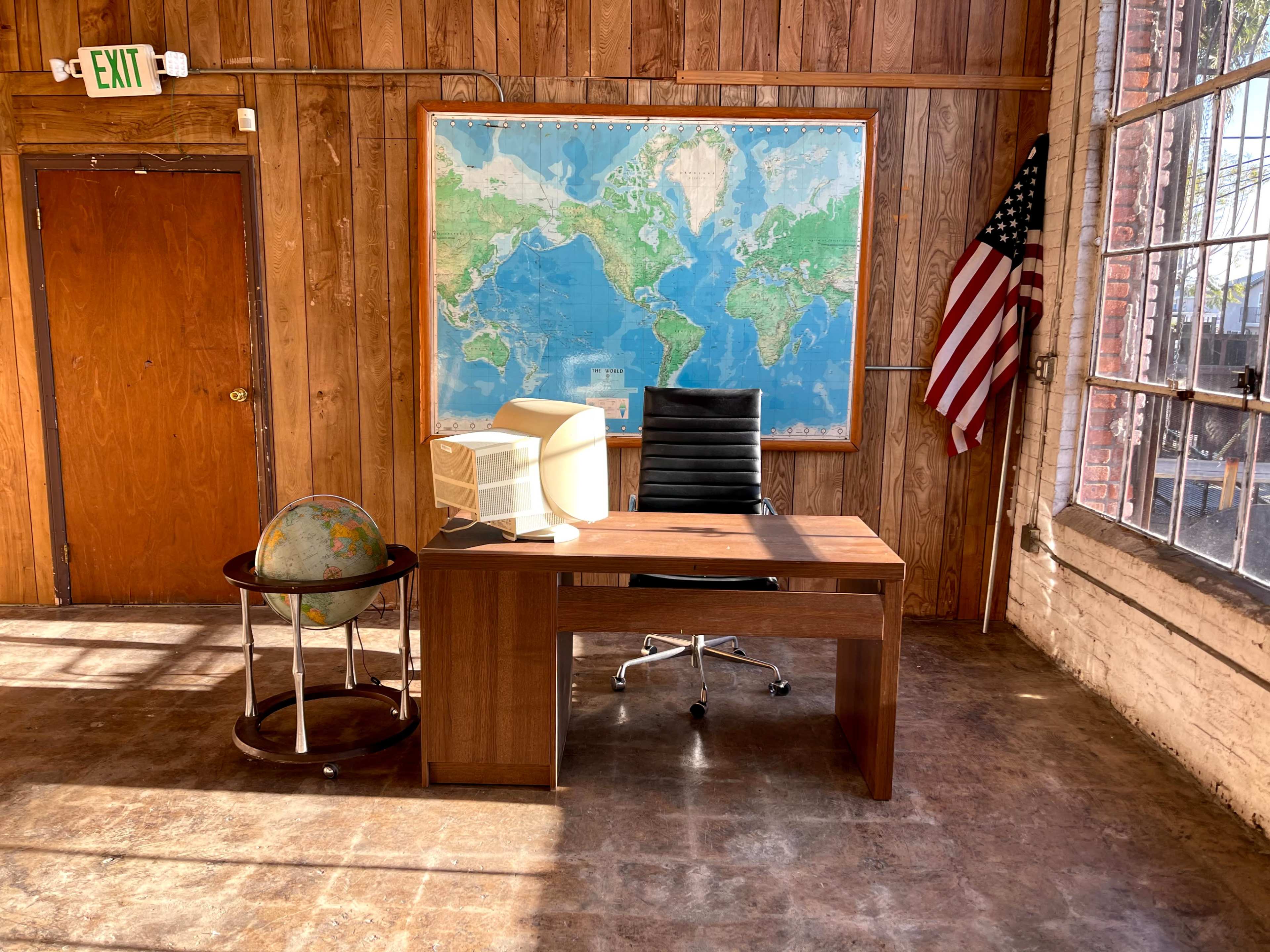 An office space features a wooden desk, a black office chair, a globe on a stand, and a large map of the world on the wall, with a window and an American flag in the background.