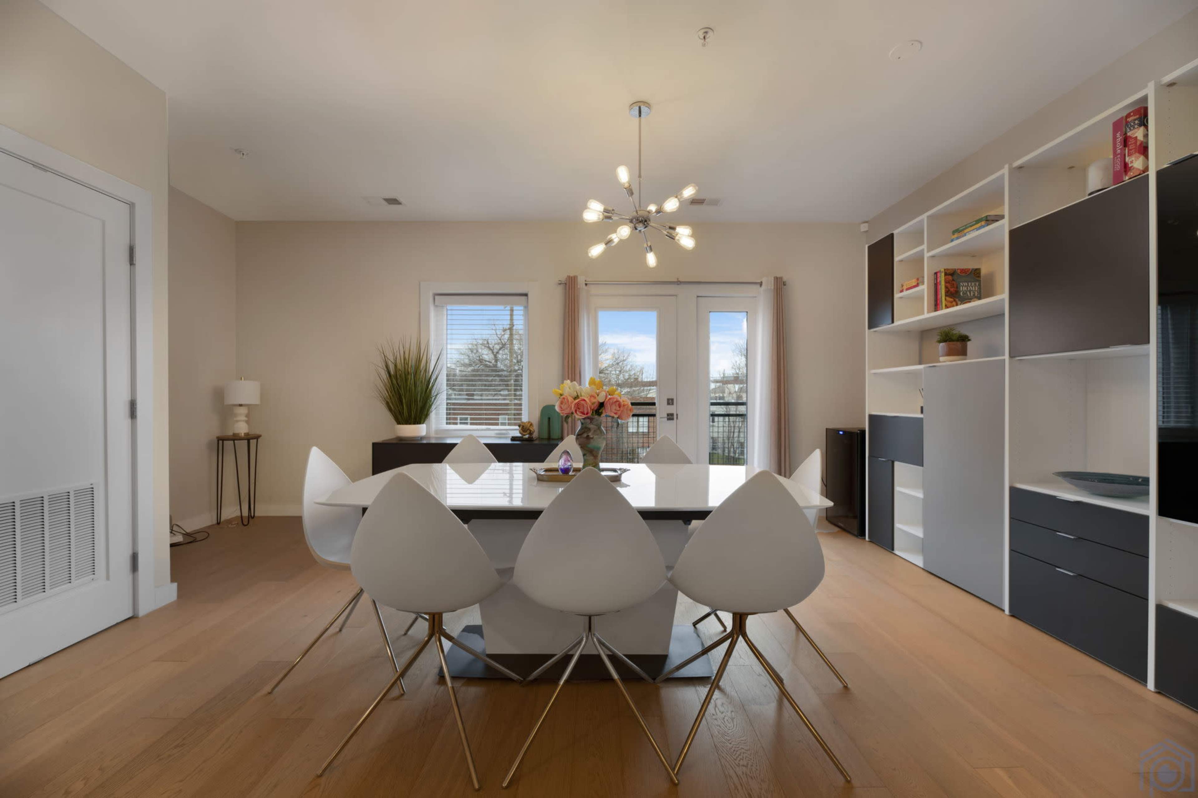 A modern dining area features a round table surrounded by white chairs, with a light fixture overhead and shelves filled with items along one wall.