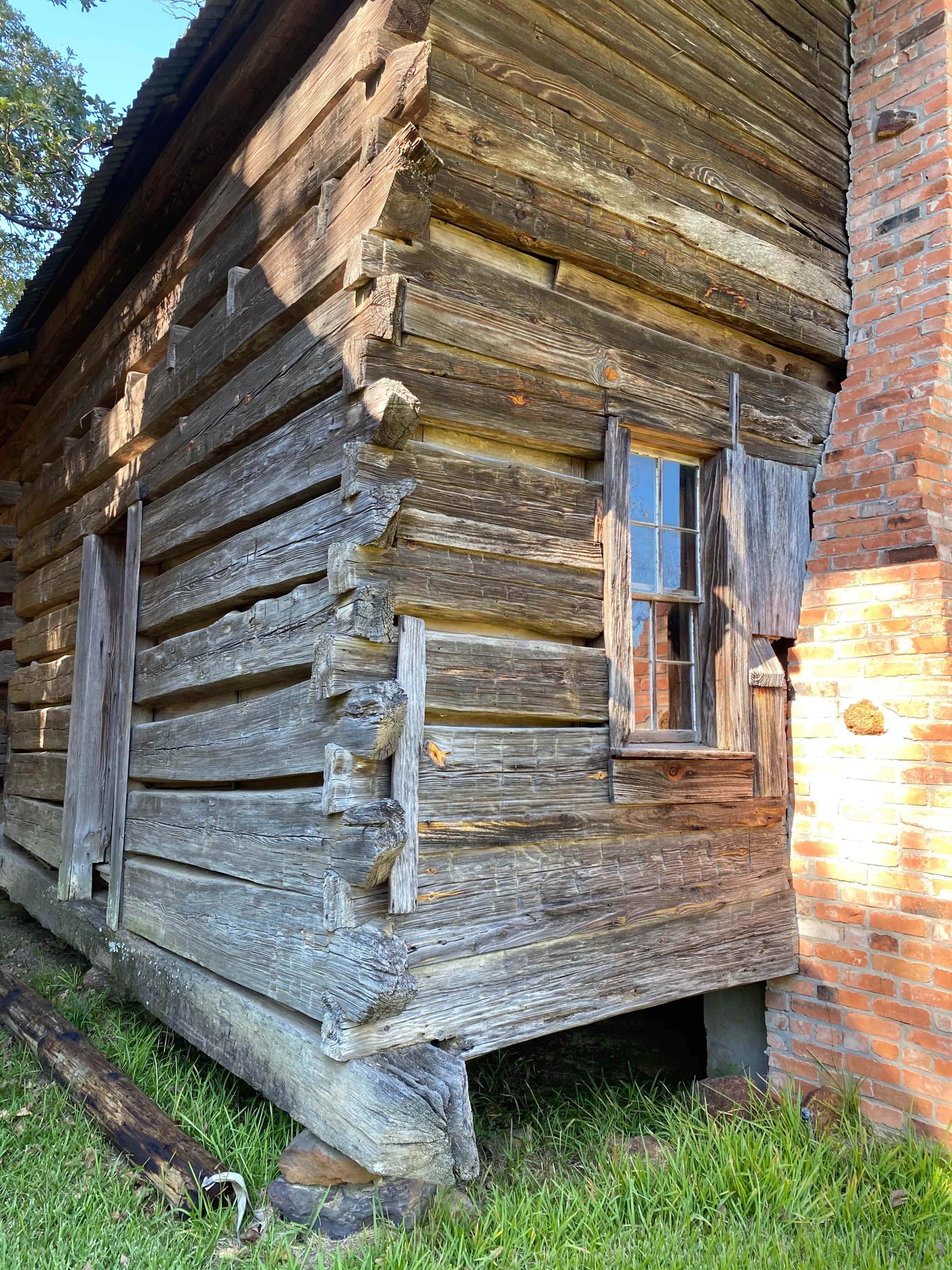 A weathered log cabin corner with a wooden window and a brick wall adjacent to it.