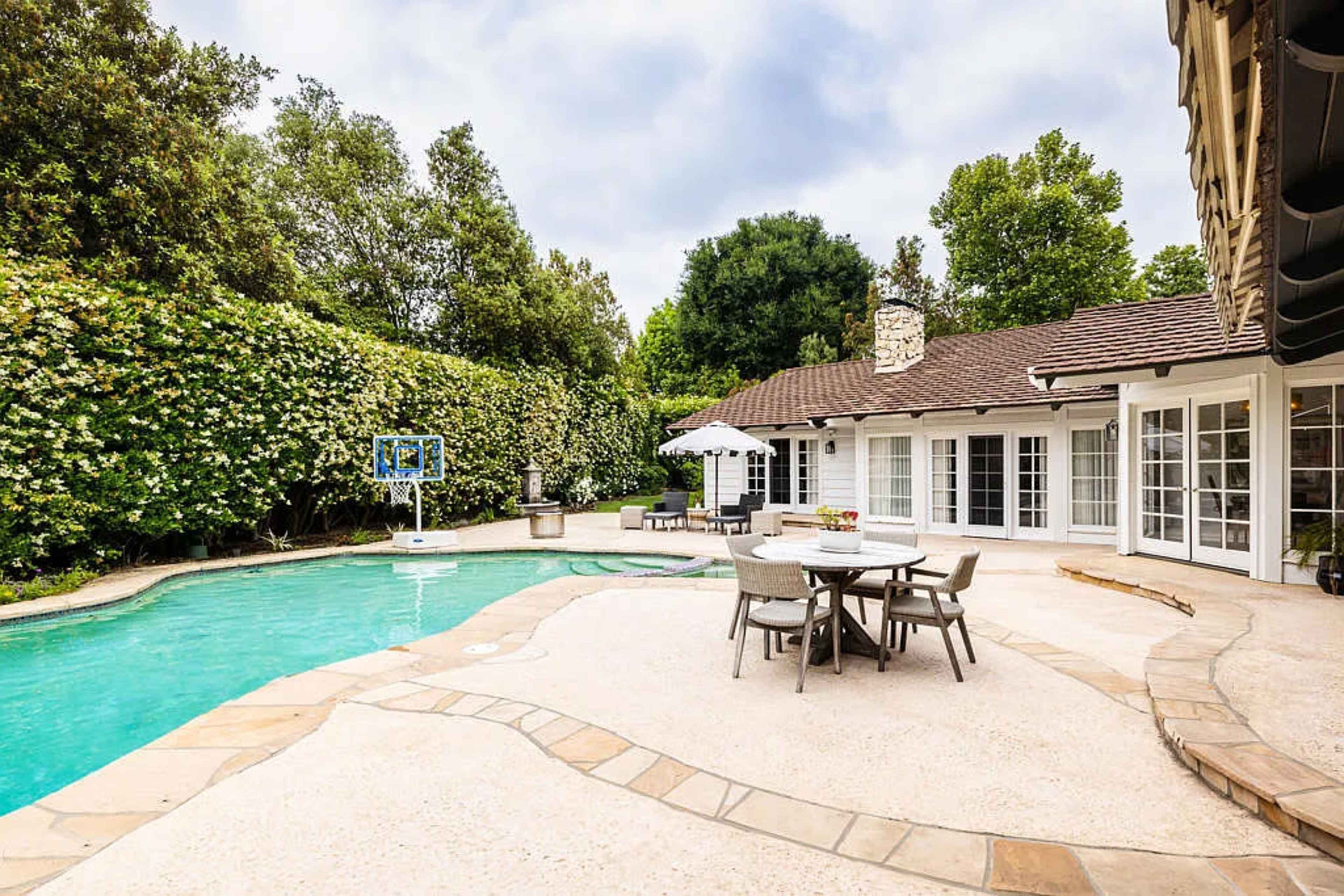 A backyard with a swimming pool, dining area, and lush greenery surrounding a house.