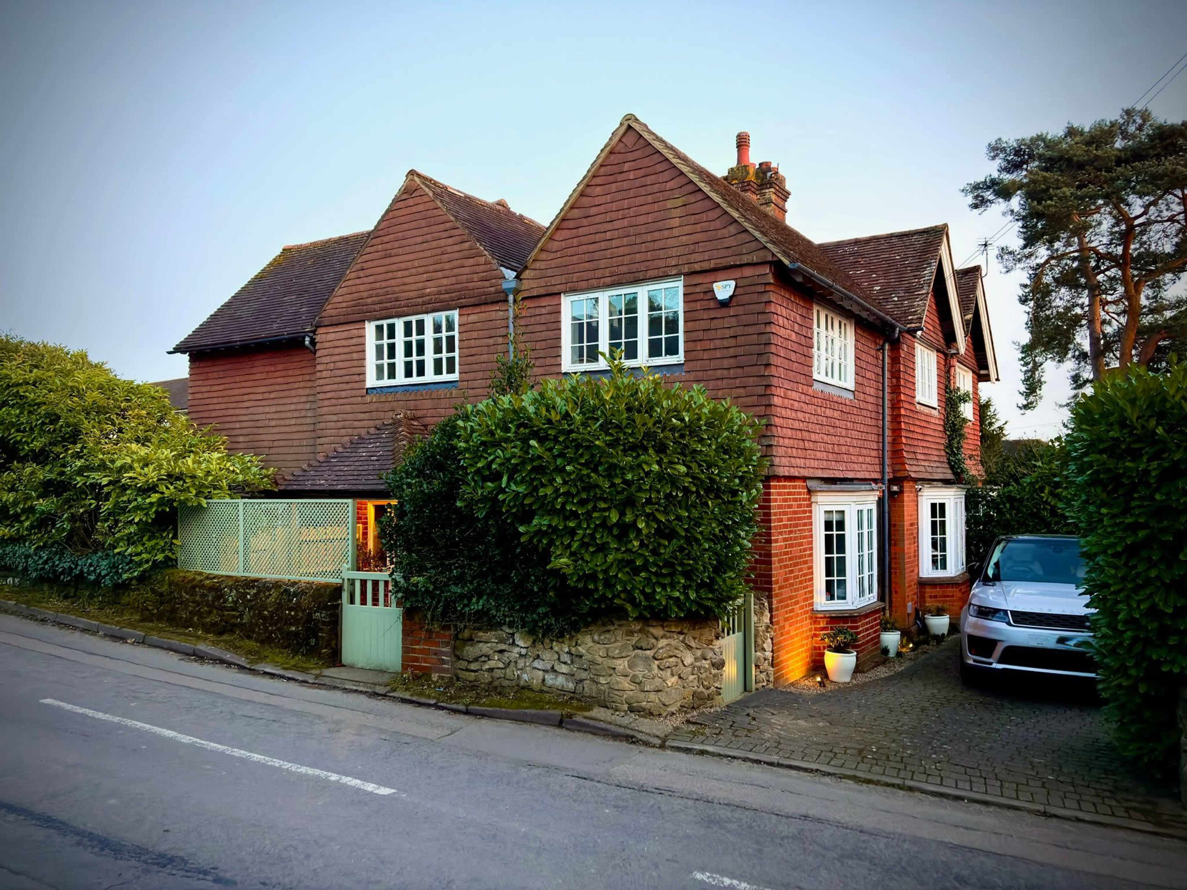 The image shows a two-story house made of red-brown wood shingles, surrounded by greenery and located beside a road with a parked car in the driveway.