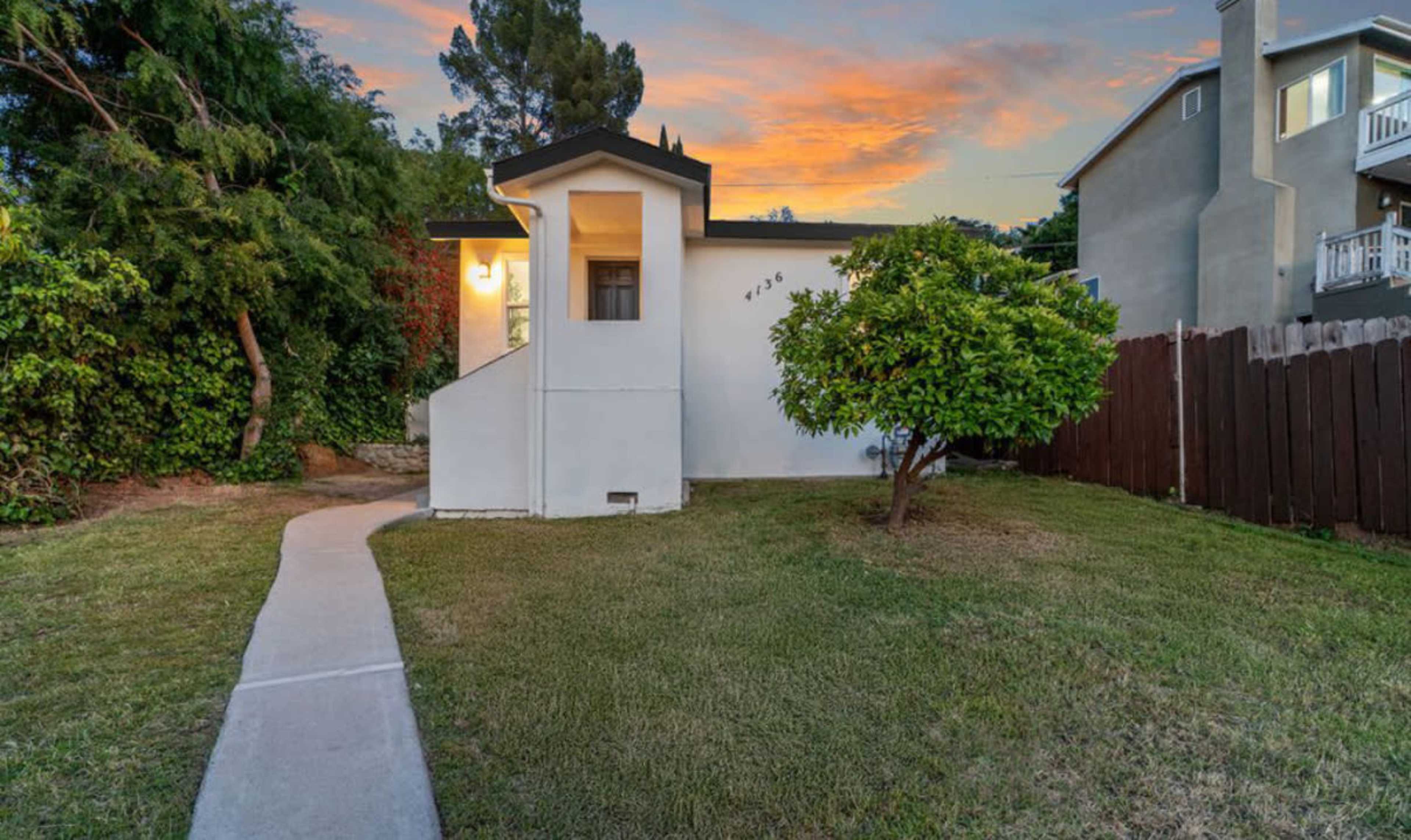 The image shows a small white house with a walkway leading up to the entrance, surrounded by green grass and trees at sunset.