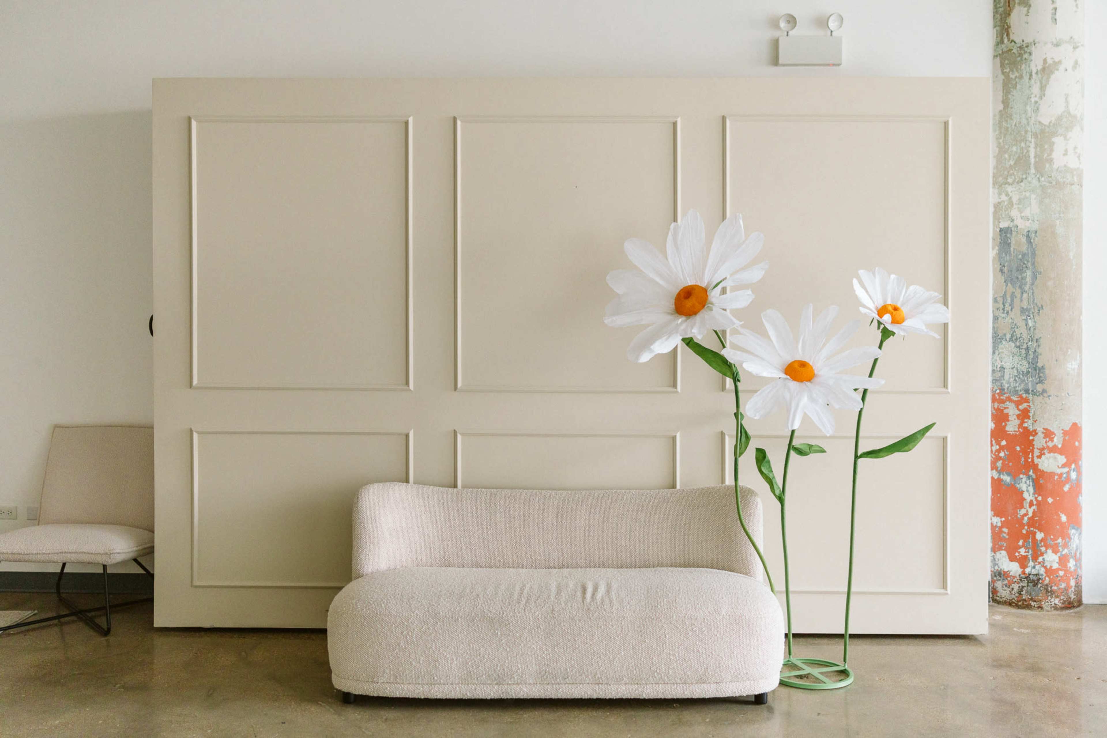 The image features a minimalist interior with a low, curved beige sofa next to large, decorative flowers on green stems set against a plain beige wall.