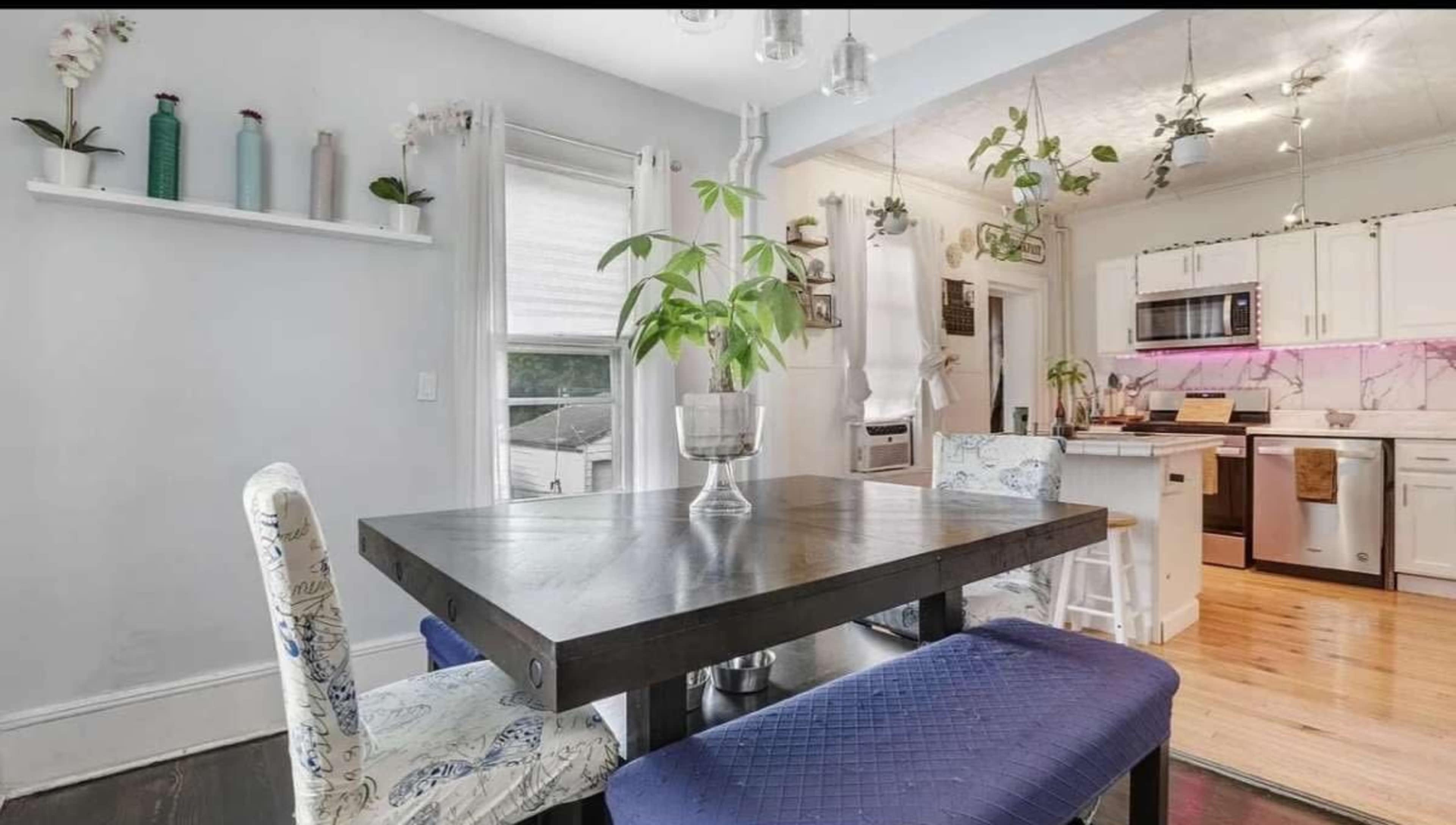 A dining area features a dark wooden table with blue upholstered chairs, surrounded by white walls and a kitchen visible in the background.