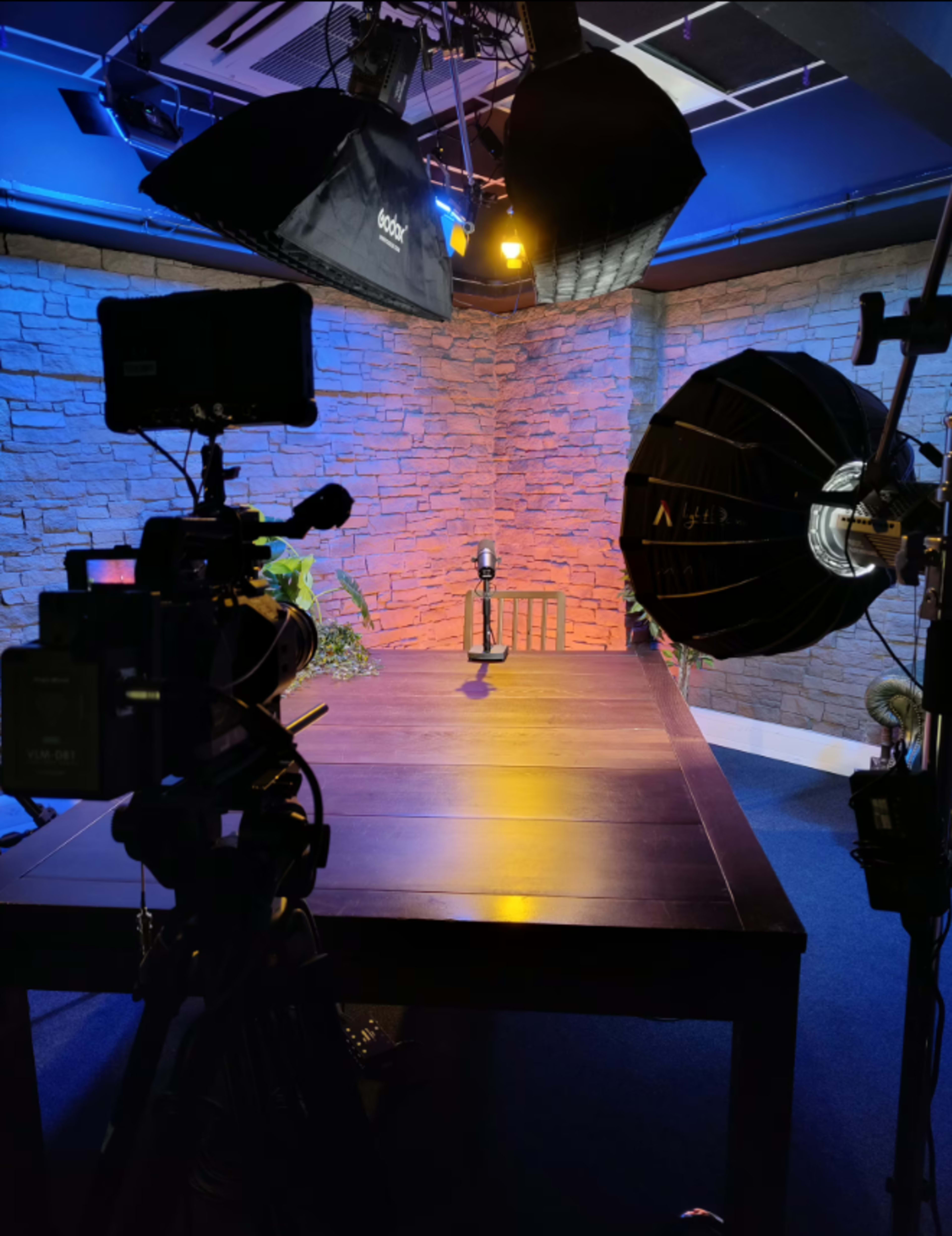 The image shows a well-lit studio setup with a microphone on a table in the center, surrounded by camera equipment and lighting in a room with stone walls.