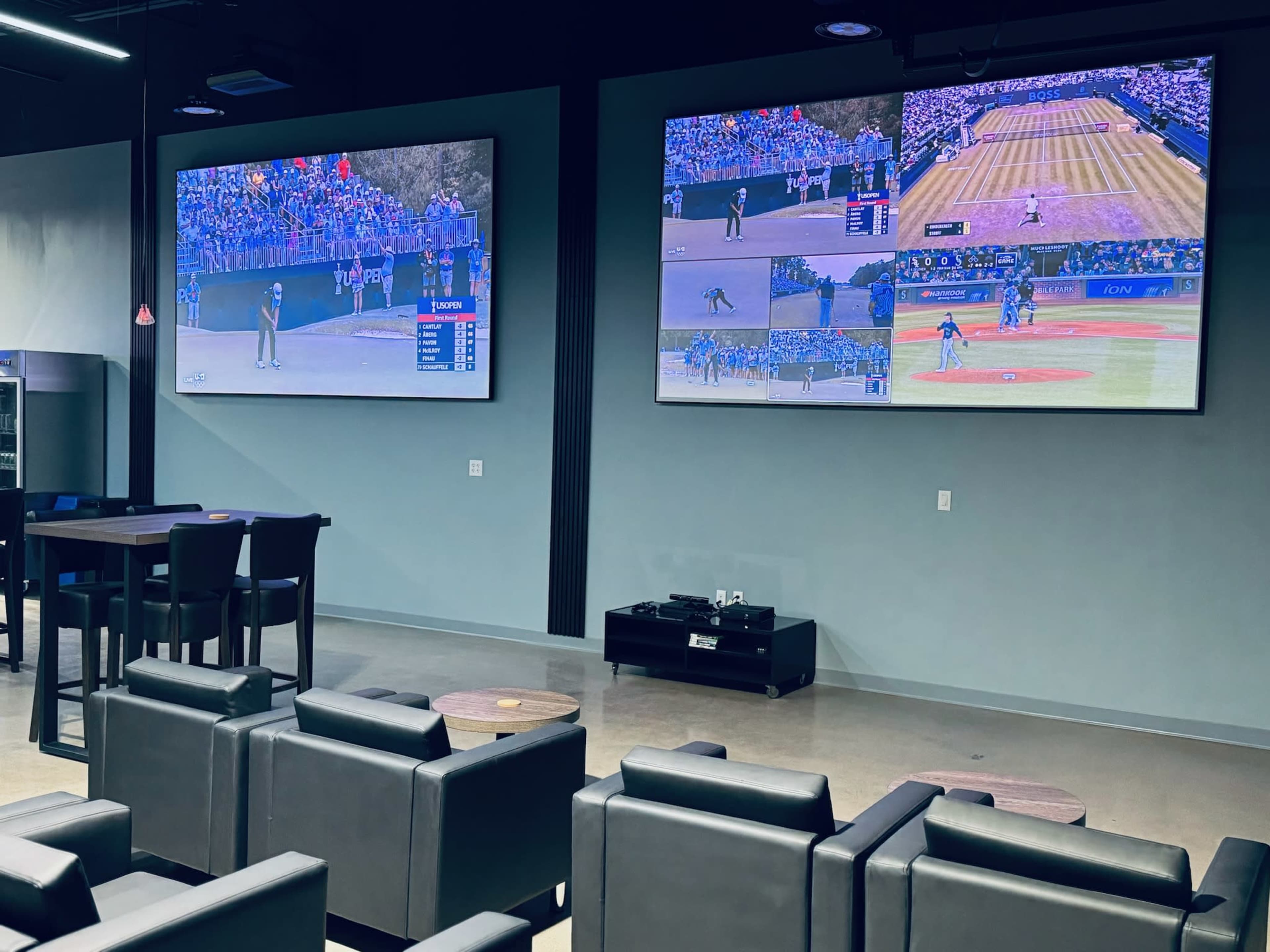 A modern room with two large screens displaying sports events, surrounded by seating arrangements consisting of black chairs and tables.