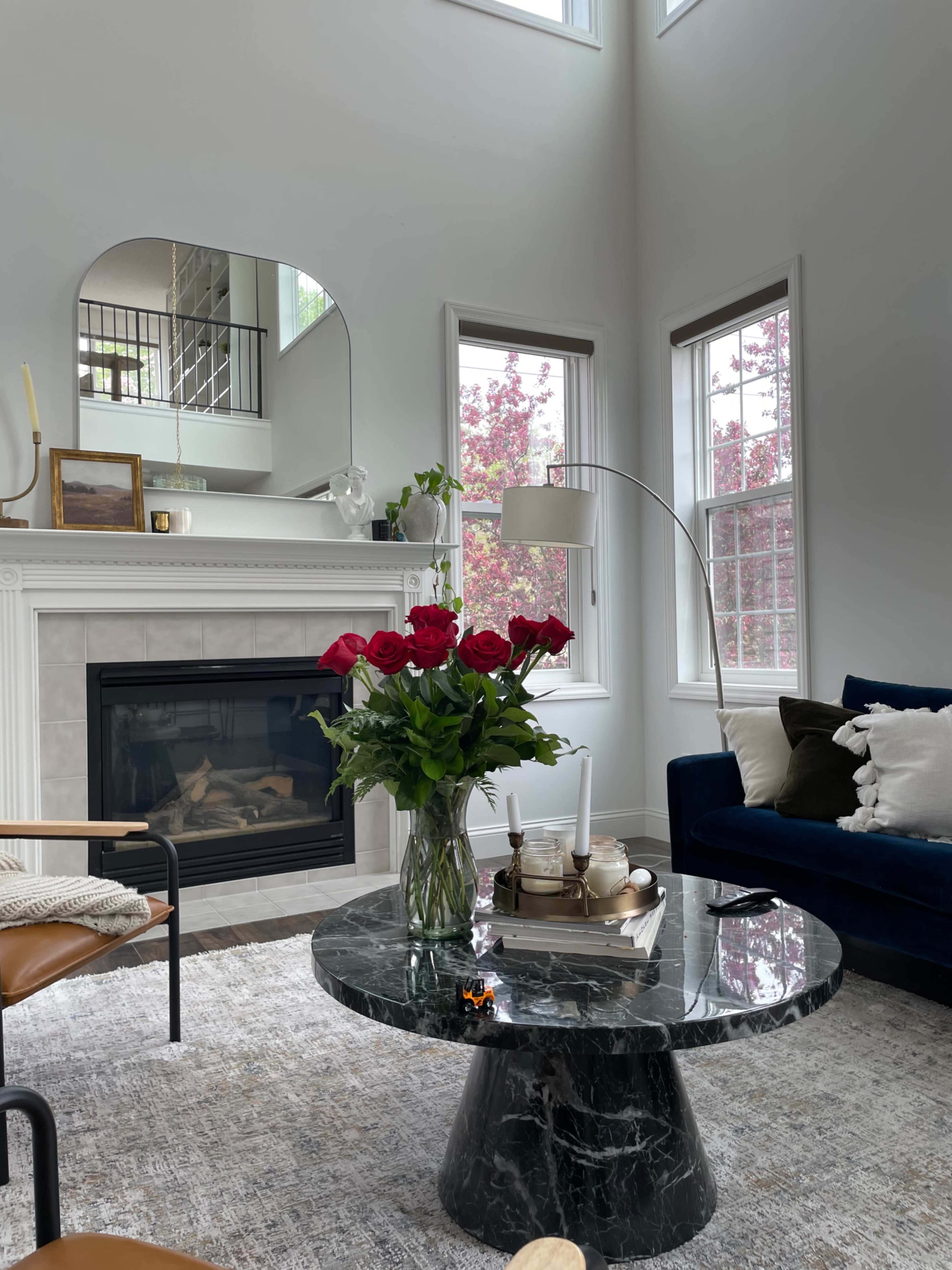 A modern living room with a marble coffee table, a fireplace, a bouquet of red roses, and large windows allowing natural light to enter.